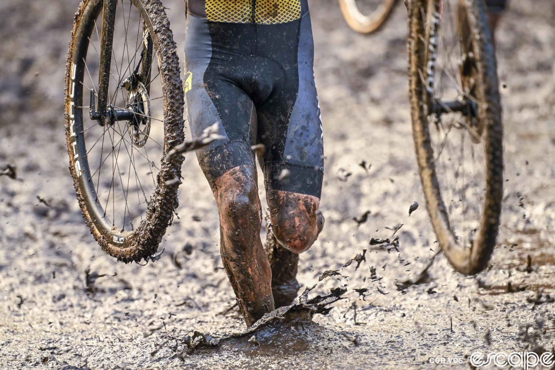 A rider is shown in closeup at the Superprestige CX in Niel. His knees and lower legs are visible with parts of two wheels as mud flies up from his footsteps.