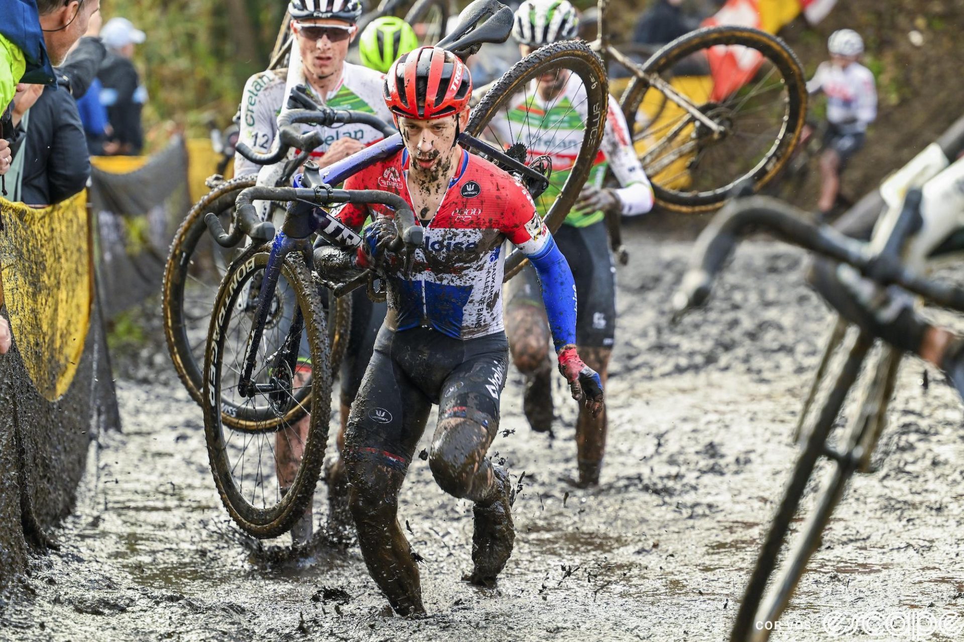 Lars van der Haar shoulders his bike to run in the mud at Superprestige Niel. He has an intense look of focus on his face.