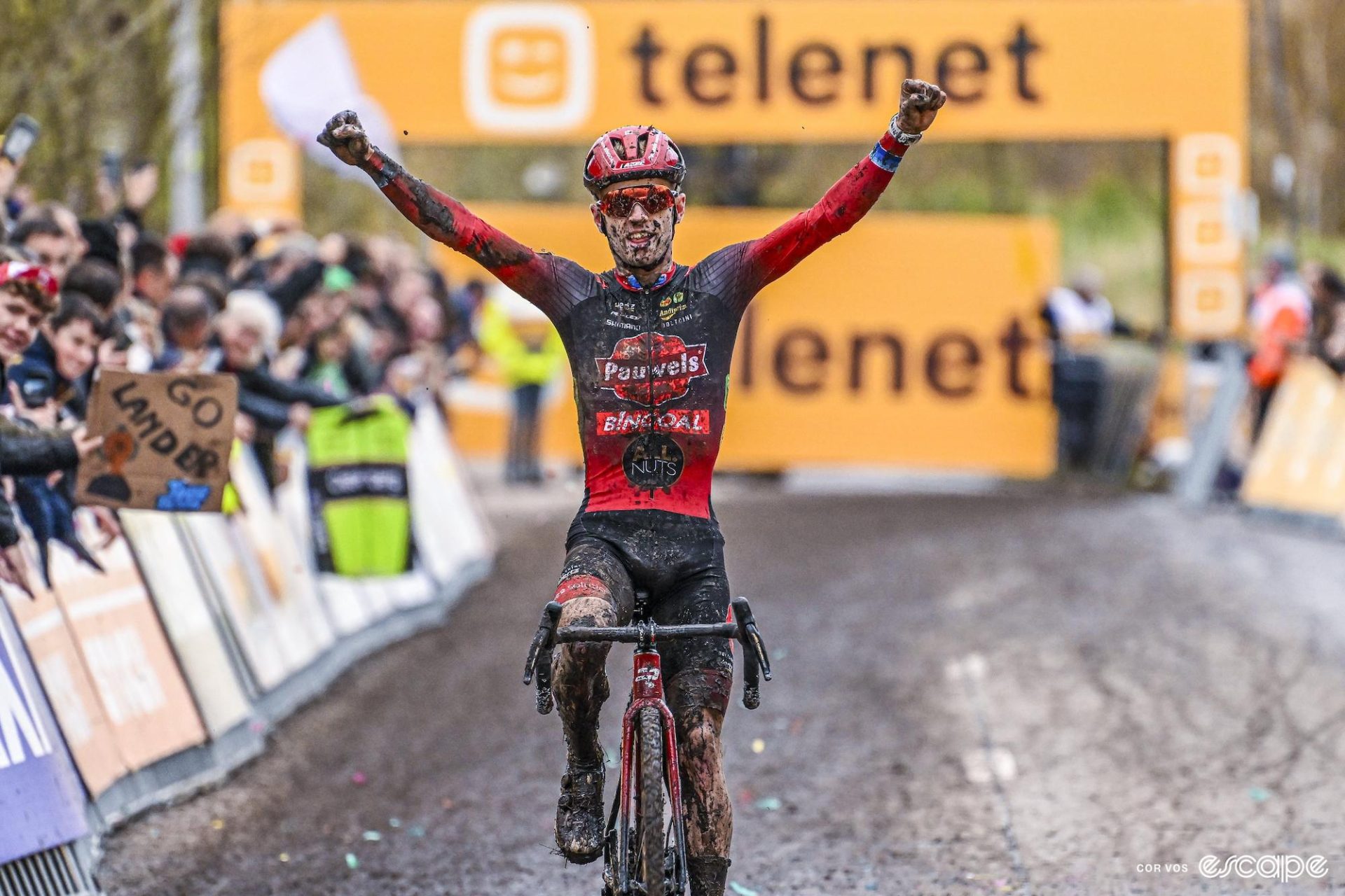 Eli Iserbyt raises his arms as he crosses the finish line to win the Superprestige Niel. He's covered in mud.