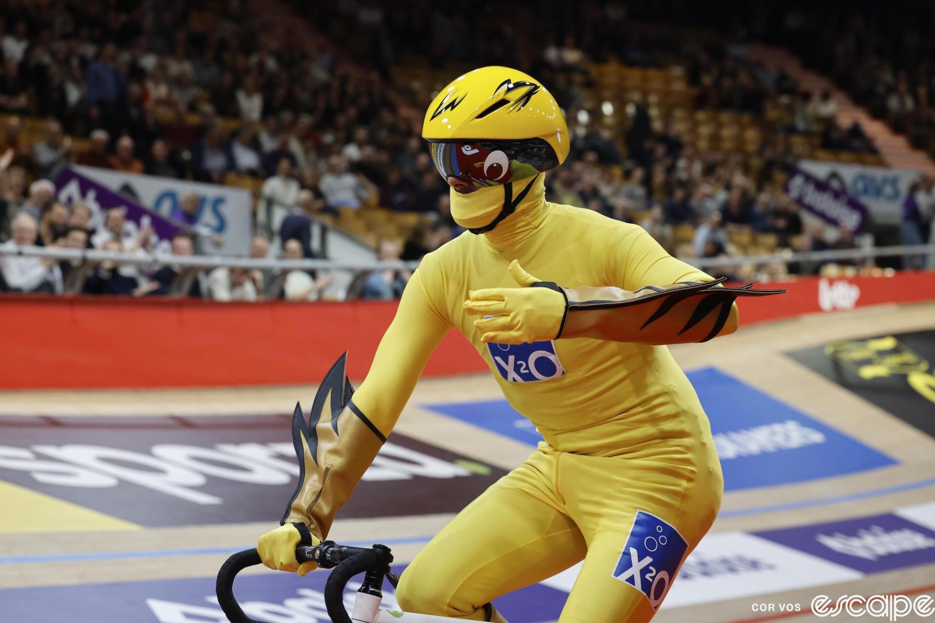 A man dressed in a full-length yellow skinsuit with stylized wrist cuffs that have flames extending off one side. His face is obscured behind a balaclava, visor, and helmet.