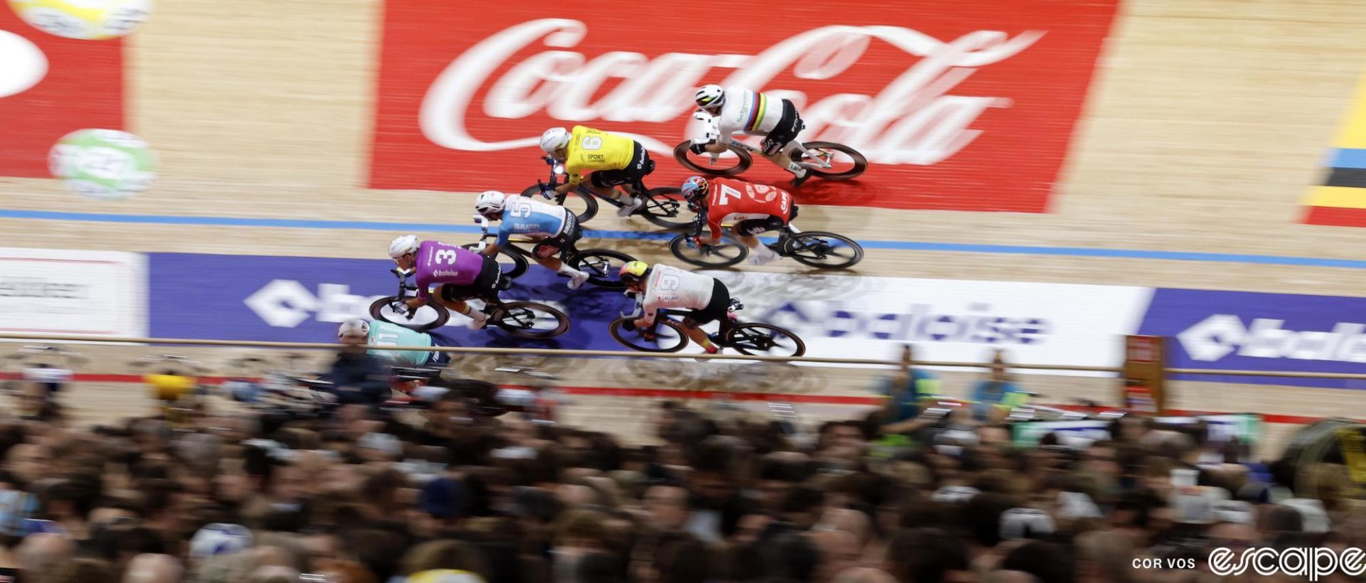 Six racers ride in a tight pack through one of the corners on the Gent track. The Coca-cola logo behind them is a blur, and the blurred infield is full of fans.