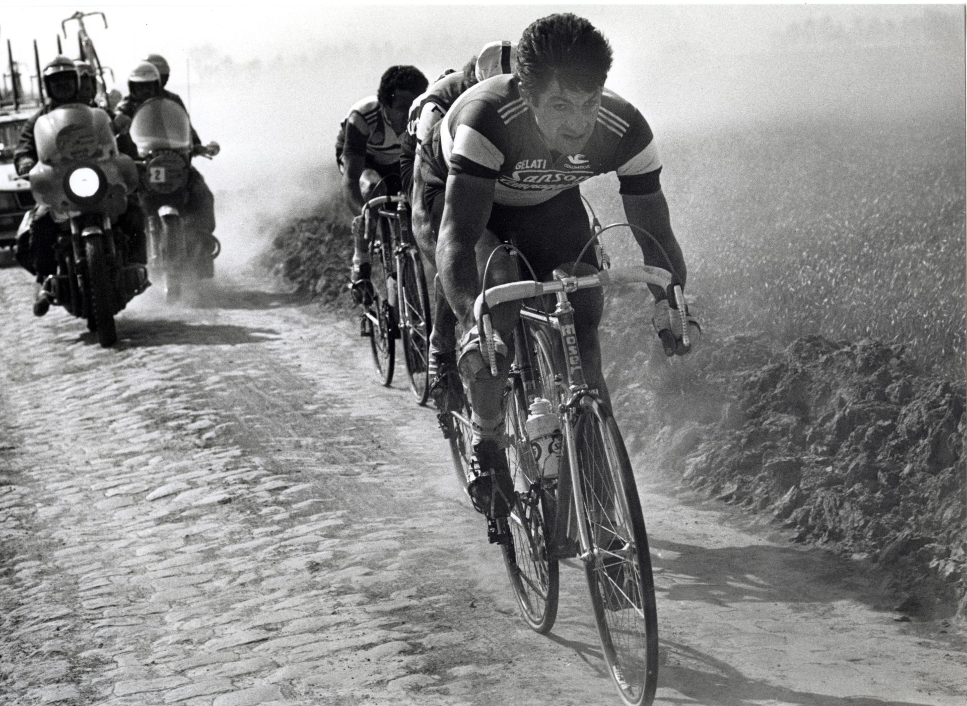 Riders race in an old edition of Paris-Roubaix, across dusty cobbles.