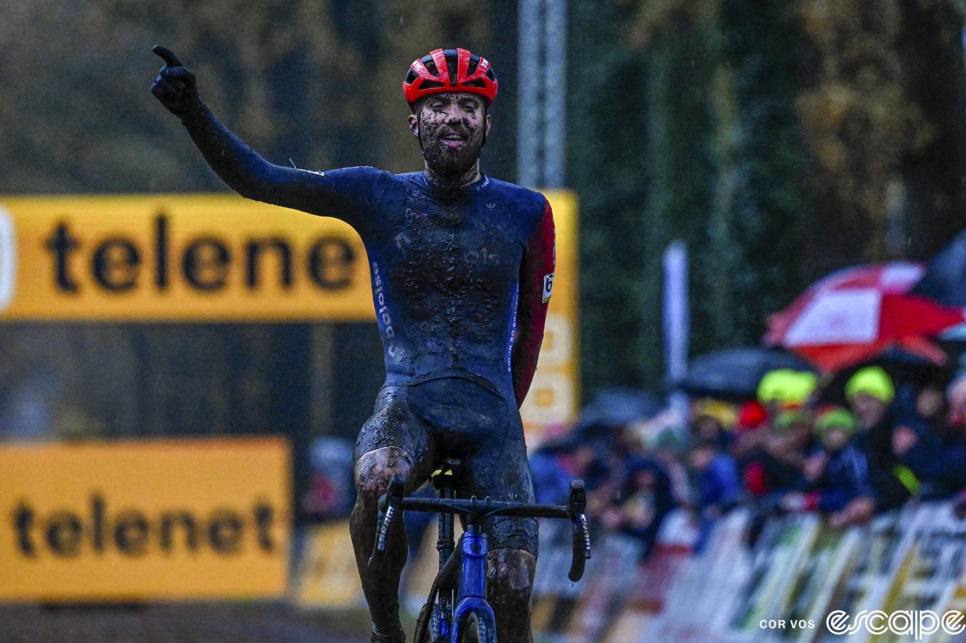 Joris Nieuwenhuis offers a one-handed victory salute as he wins Superprestige Merksplas. His blue kit is covered in mud, logos unreadable, and his full beard is also full of mud up to around his eyes.