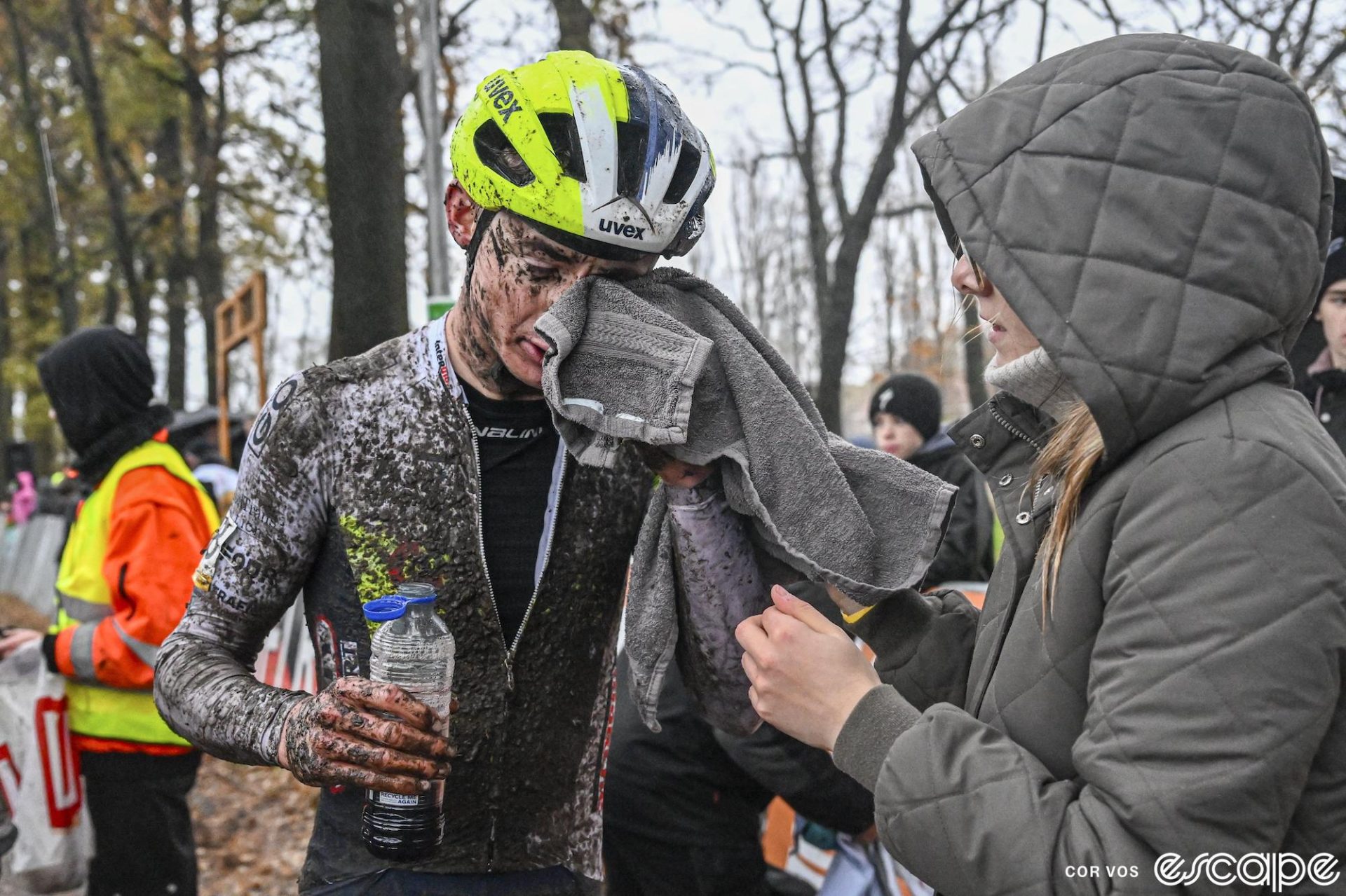 Gerben Kuypers wipes mud from his face with a towel handed to him by a woman. He holds a water bottle in front of his filthy, unzipped skinsuit.