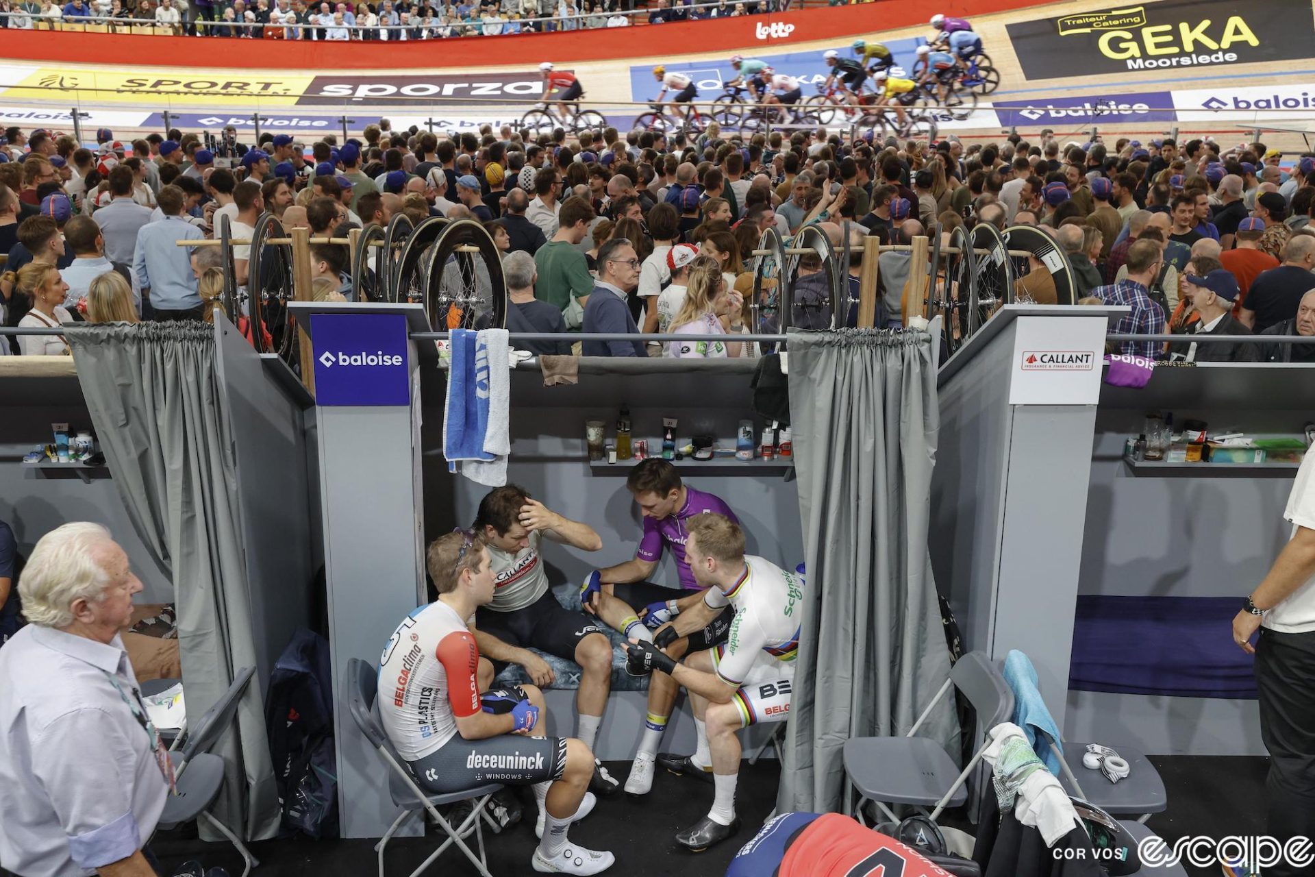 Four racers sit in a circle in an infield cabin, two on the bed and two on folding chairs. Behind them, the race continues with their teammates.