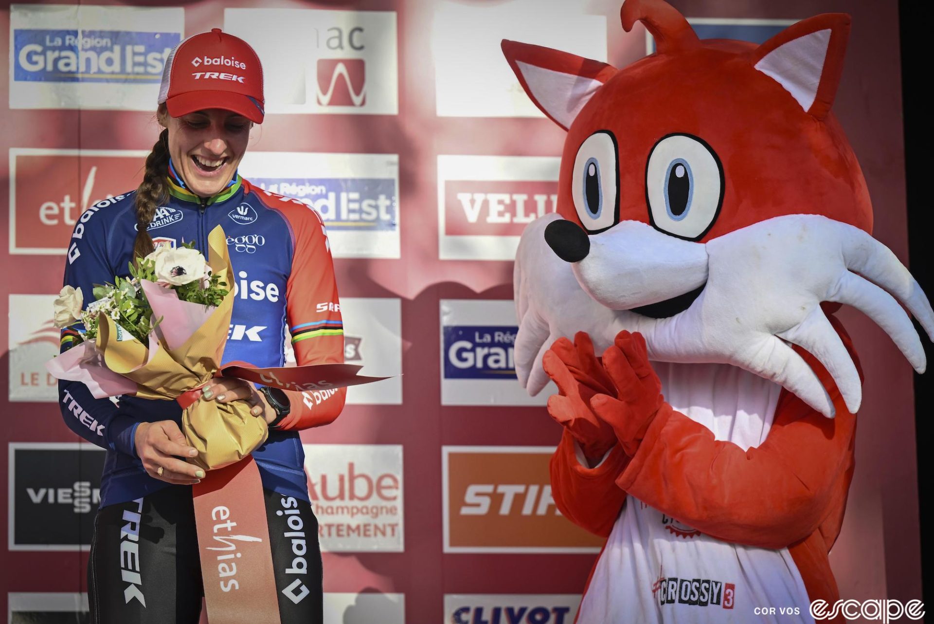 Lucinda Brand receives flowers on the podium from a person in a mascot costume that looks to be a large red fox with white face and belly.