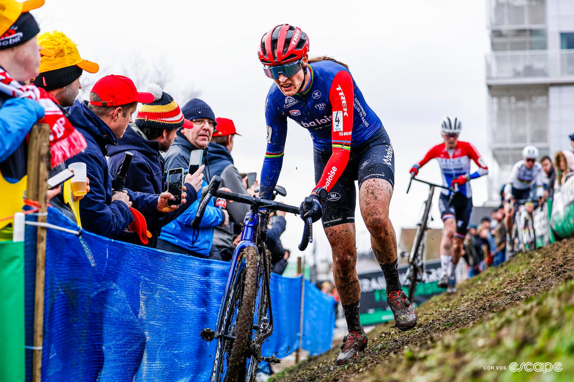 Mud covering her legs, Lucinda Brand grimaces as she pushes her bike through the off-camber section during X20 Trofee Kortrijk, watched by fans wrapped up agianst the cold.