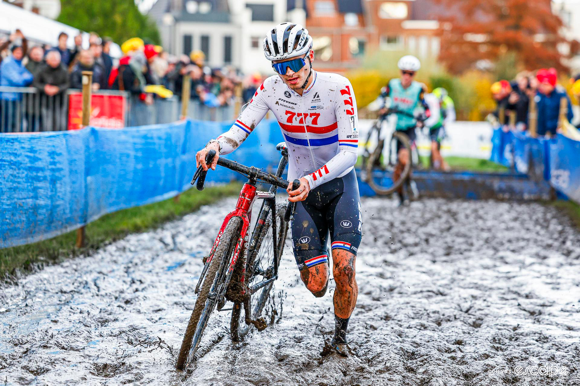 British national champion Cameron Mason splashes through thick, wet mud, running with his bike, at X20 Trofee Kortrijk, riders seen leaping the boards in the background.