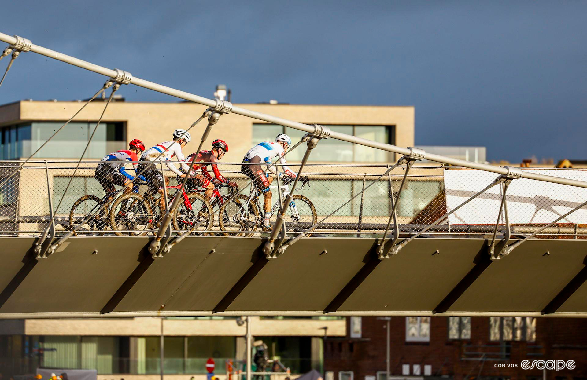 The four leaders of the men's cyclocross X20 Trofee Kortrijk ride a flyover as a group, lit up late in the day against a gloomy backdrop.