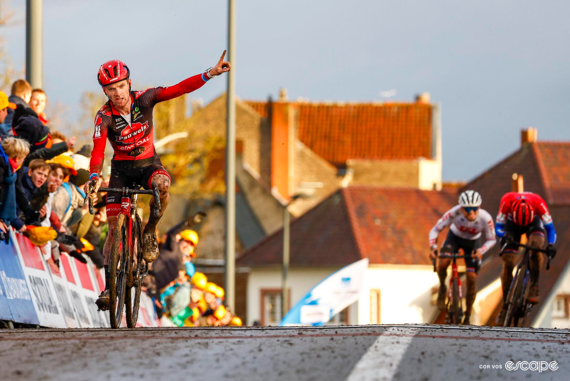 With the sun almost set, Eli Iserbyt reaches the finish line with one finger pointed skyward in celebration as Lars van der Haar and Cameron Mason sprint for second just behind.
