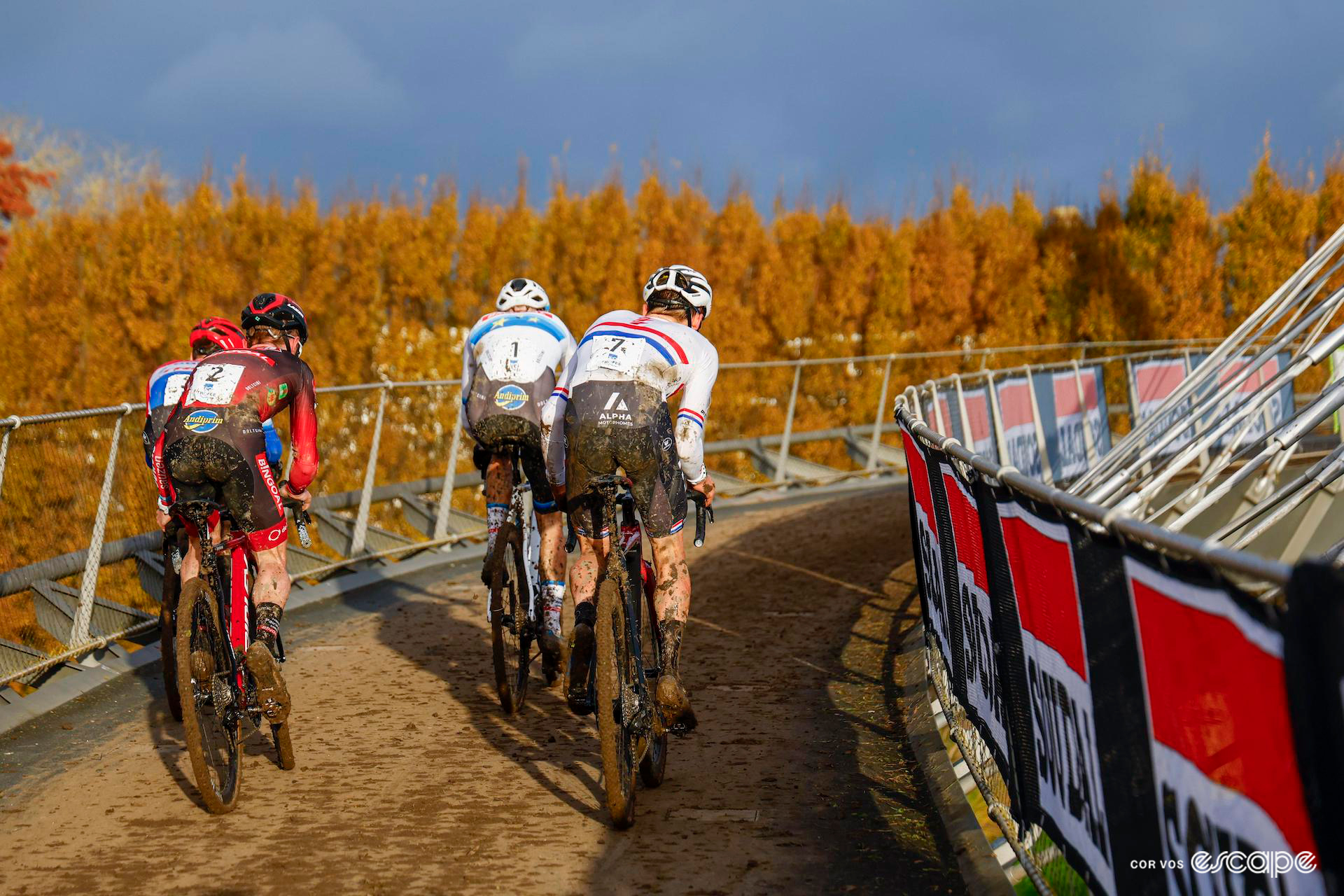 The four leaders of the men's cyclocross X20 Trofee Kortrijk seen from behind as they ride onto a muddy flyover as a group, lit up late in the day against an autumnal backdrop.
