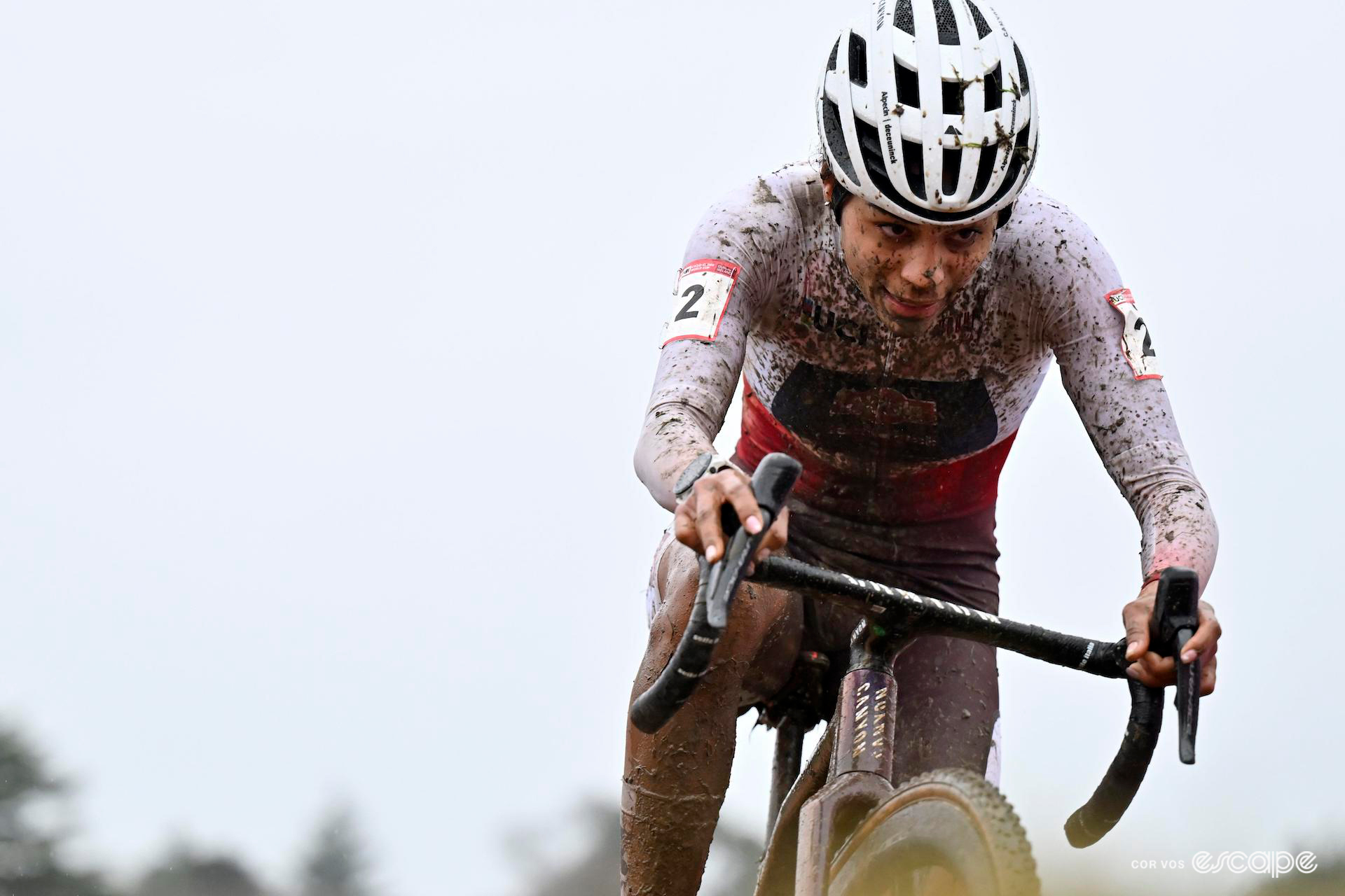 Medium shot of Ceylin del Carmen Alvarado, her white and red World Cup leader's jersey is splattered with mud, digs deep during CX World Cup Dublin, an overcast sky overhead.