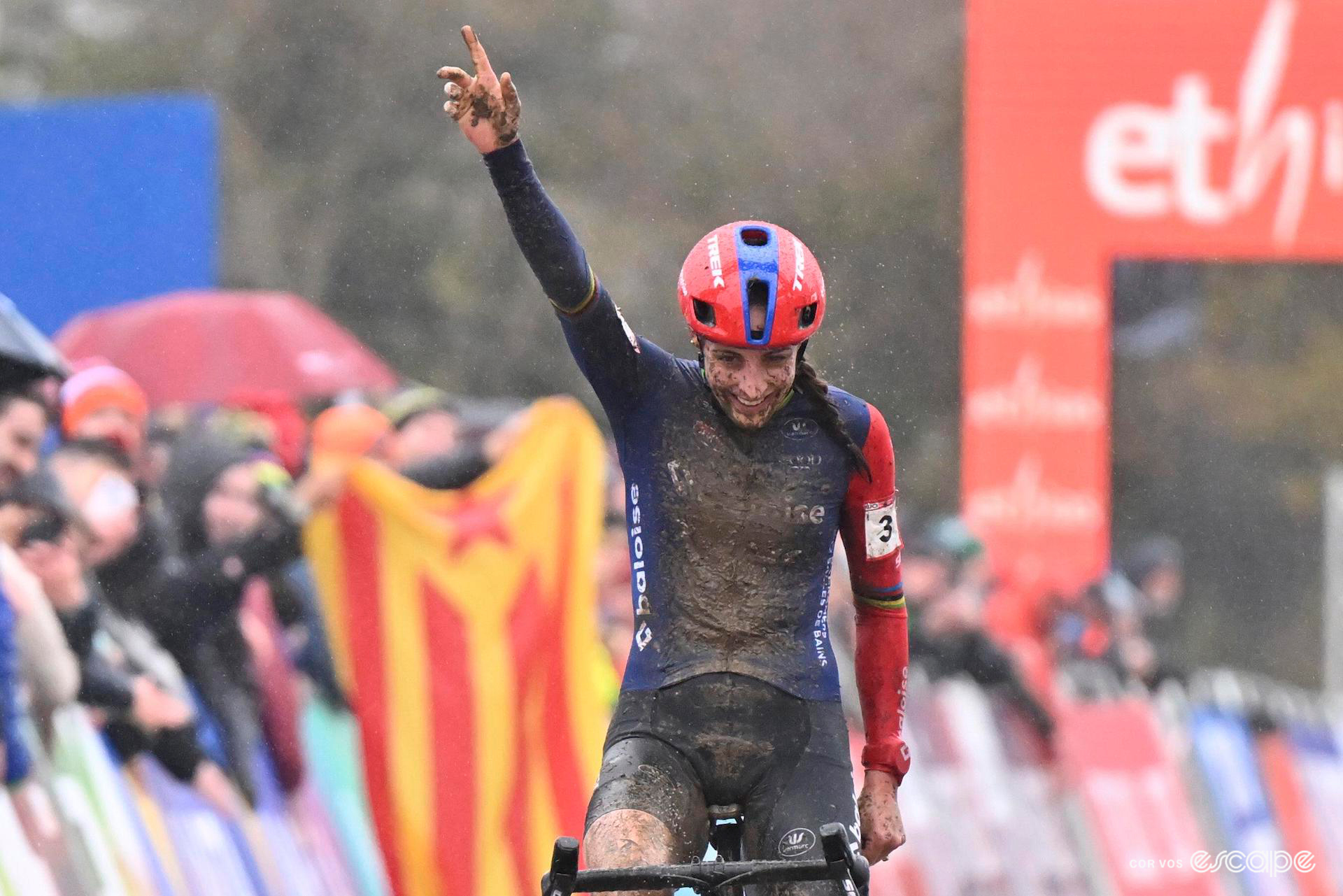 Medium shot of Lucinda Brand of Baloise Trek Lions, the front of her jersey caked in dark mud, as she smiles to herself and points one finger to the sky to celebrate victory at CX World Cup Dublin.