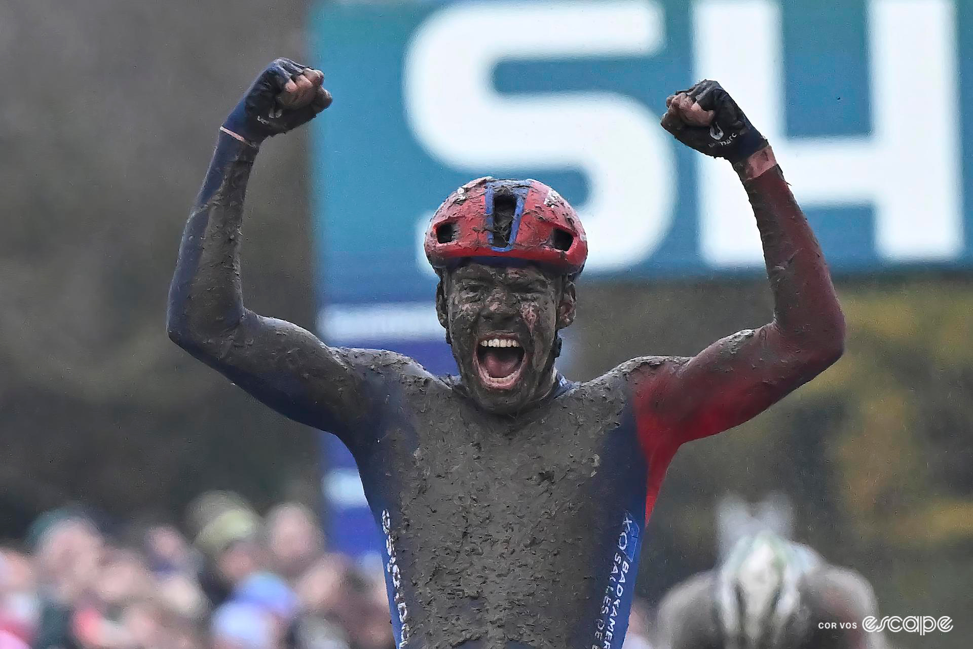 Medium shot of Pim Ronhaar of Baloise Trek Lions, the front of his jersey and face caked in dark mud, as he raises both fists in the air and roars to celebrate CX World Cup Dublin victory over Laurens Sweeck, whose head is bowed in the background.