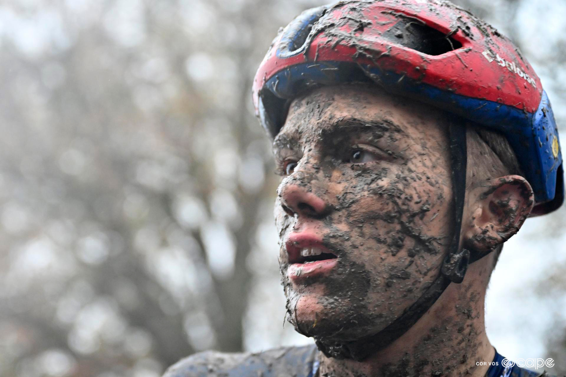Close-up of Thibau Nys after CX World Cup Dublin, his face and helmet splattered with mud at various degrees of dryness, gazing off into the distance after a hard race.