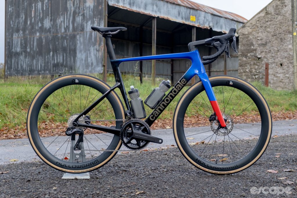 The photo shows a new Cannondale SuperSix Evo Hi-Mod 2 with Ultegra Di2 groupset, HollowGram R-SL 50 wheelset, Prologo saddle, Vision Trimax Carbon Aero handlebars and Conceal stem along a country lane in front of a field and an old barn and stone gable wall in dark wintery conditions.