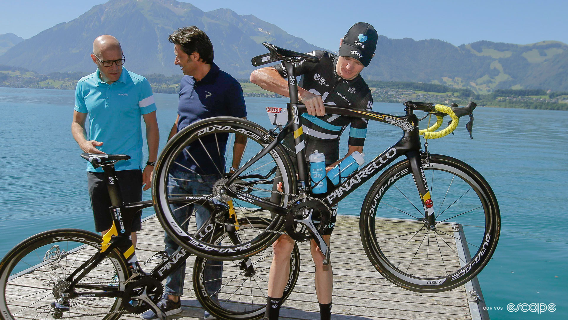 The photo shows Chris Froome standing on a small wooden boat dock holding up his Pinarello F8 with Sir Dave Brailsford and Fausto Pinarello to his right.