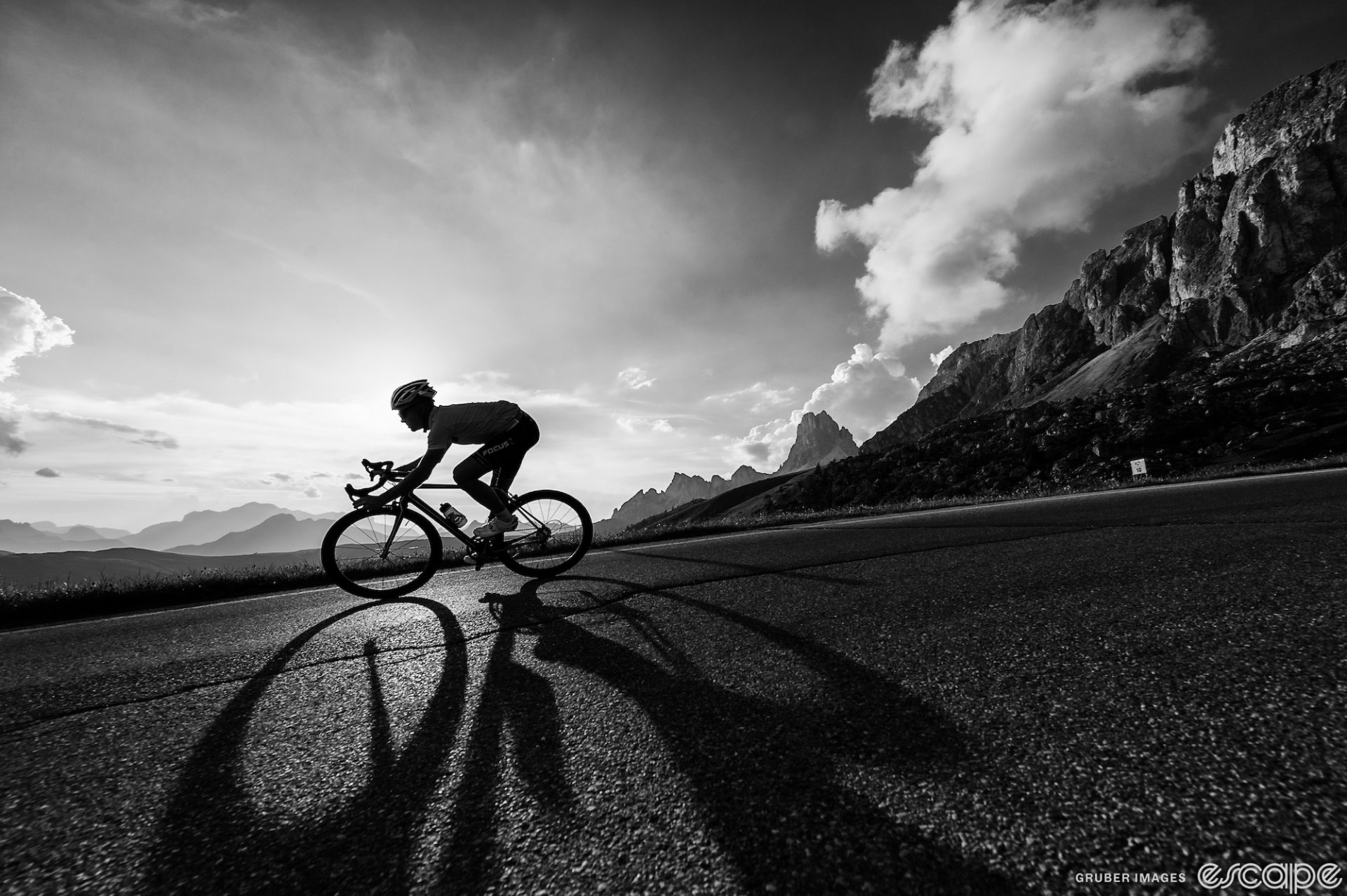 A black and white photo of a rider descending a paved road with rocky mountains in the background. The rider is backlit and silhouetted against the sky.