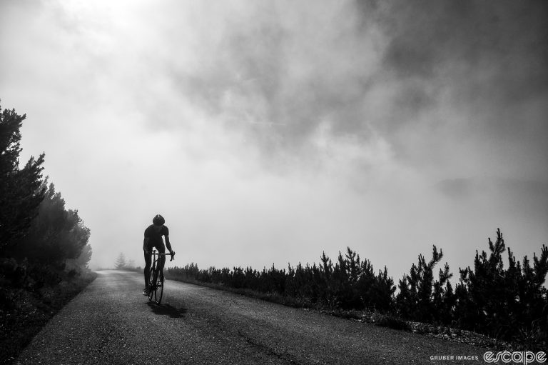 A rider climbs a road alone, out of a foggy mist in the background. The black and white shot is backlit, with the rider silhouetted against a dark road and white cloud.