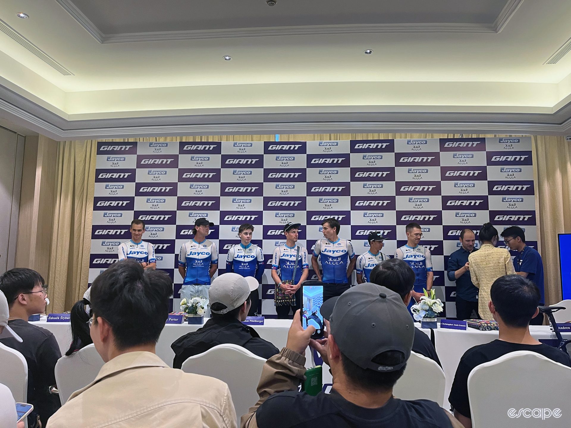 The press conference with Jayco-AlUla. Riders in team kit stand before a crowd of PR guests.