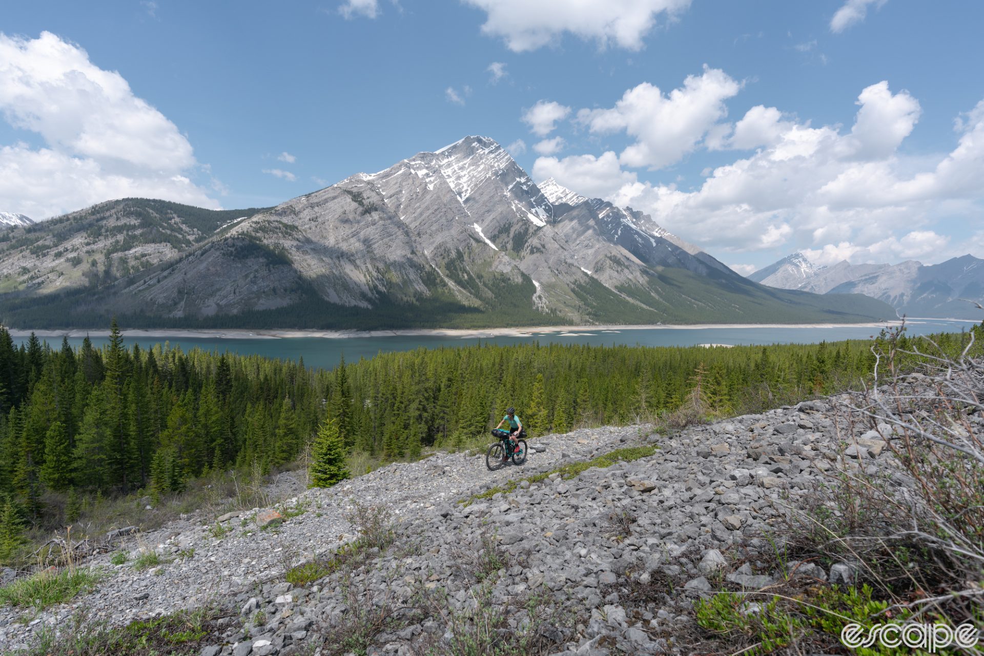 A lone cyclist traverses a loose, rocky trail above an alpine lake. A high peak in the background still has snowfields.