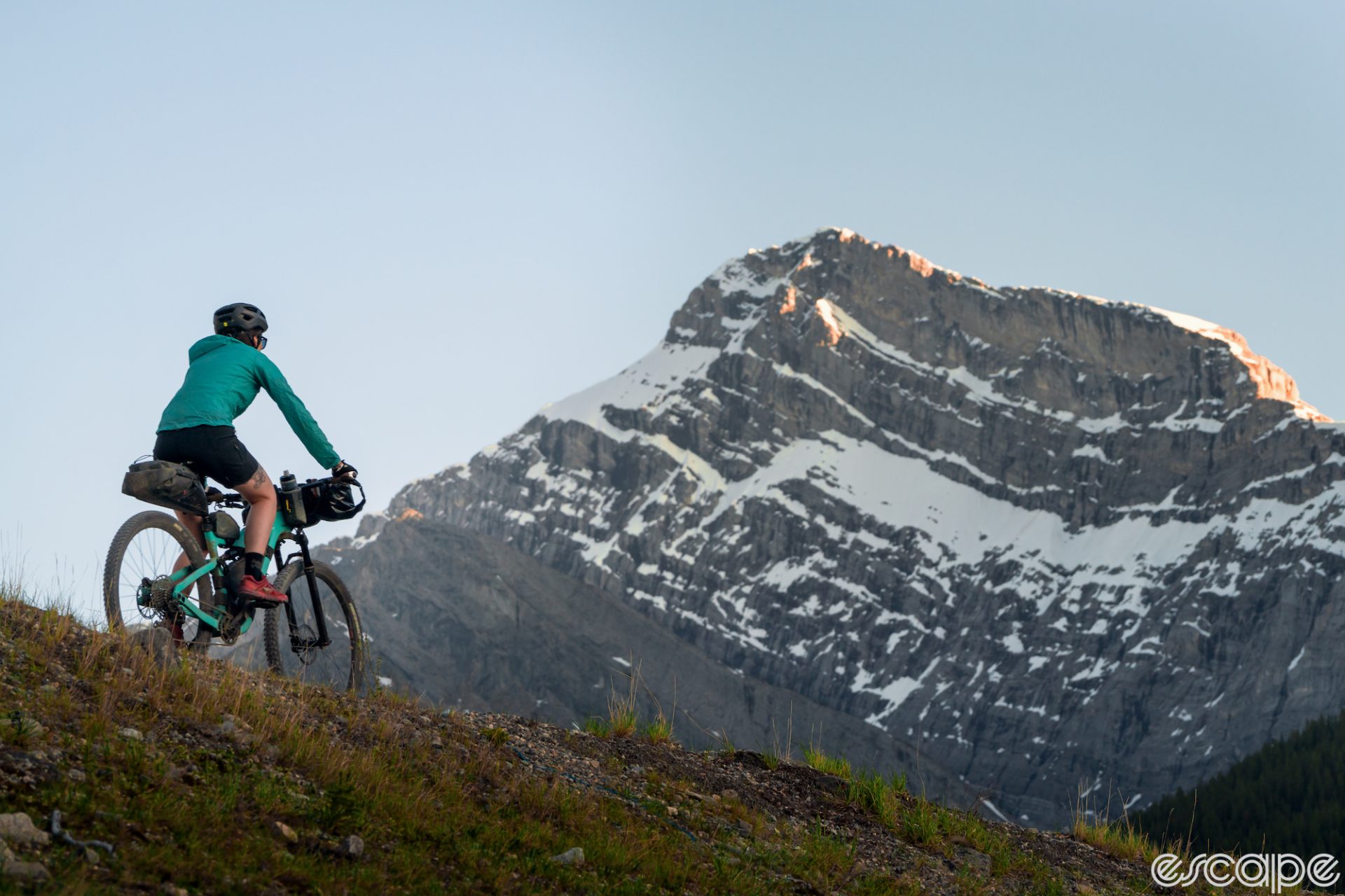 A bikepacker rides on a high alpine trail with a rugged, snow-capped peak in the distance.