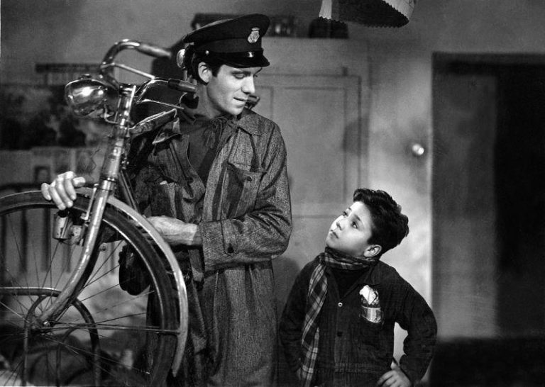 Still from 1948 Italian film Bicycle Thieves, showing main character Antonio dressed for work with his bicycle on his right shoulder, looking down out his young son Bruno.