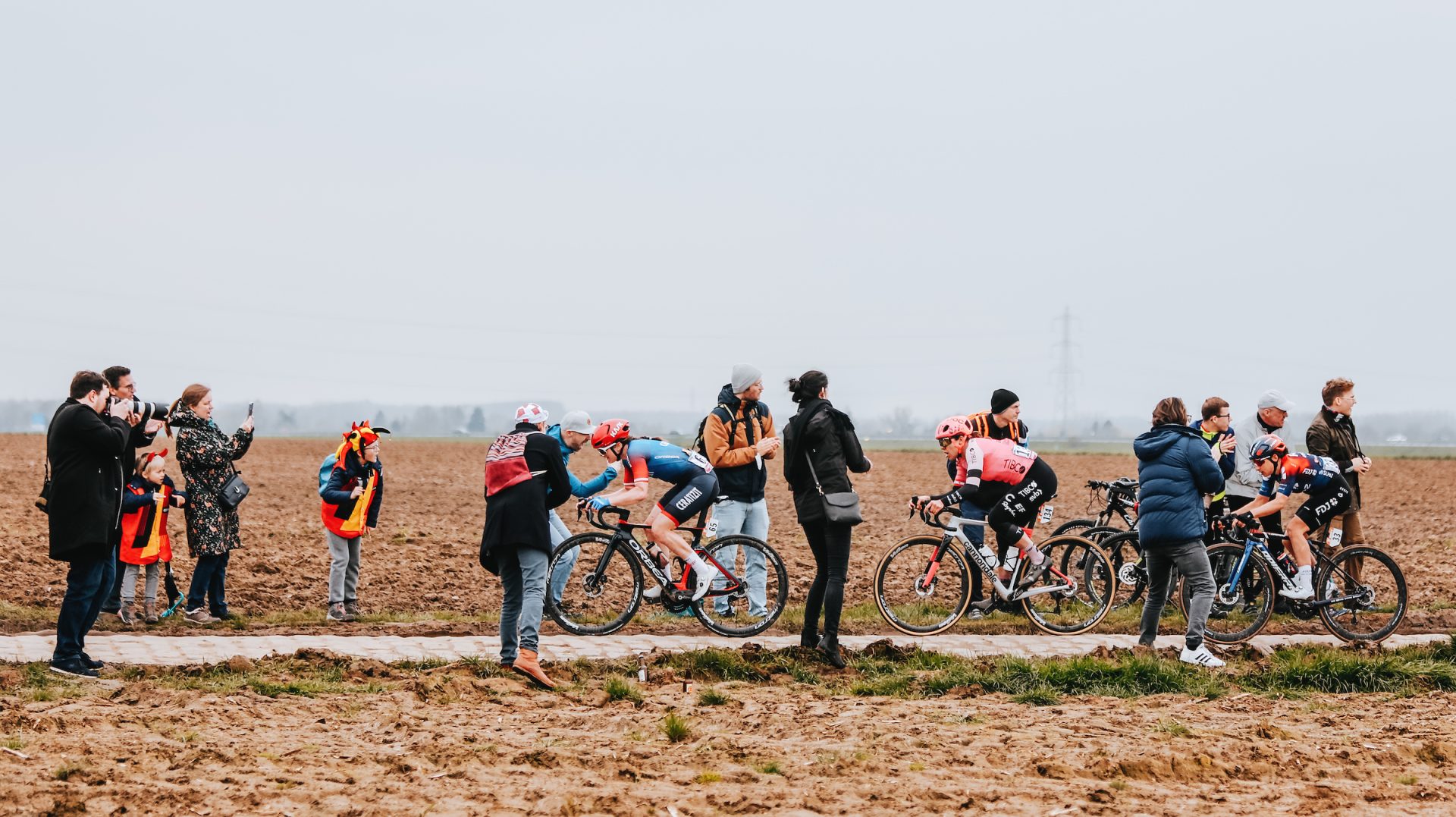Panoramic perspective of fans and photographers gathered at a narrow cobbled section at the 2023 Paris Roubaix Femmes. A string of riders fly past, navigating the slick cobbles on an overcast day in April.