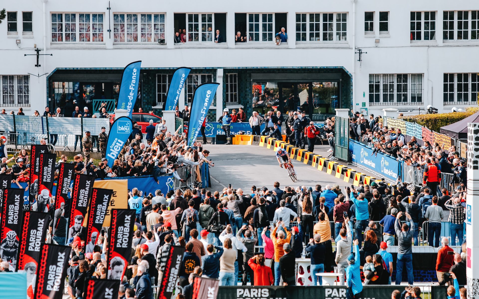 A wide shot of Mathieu van der Peol as he enters the Roubaix Velodrome for the finish. A sea of fans and sponsor banners flank the barriers of the road.