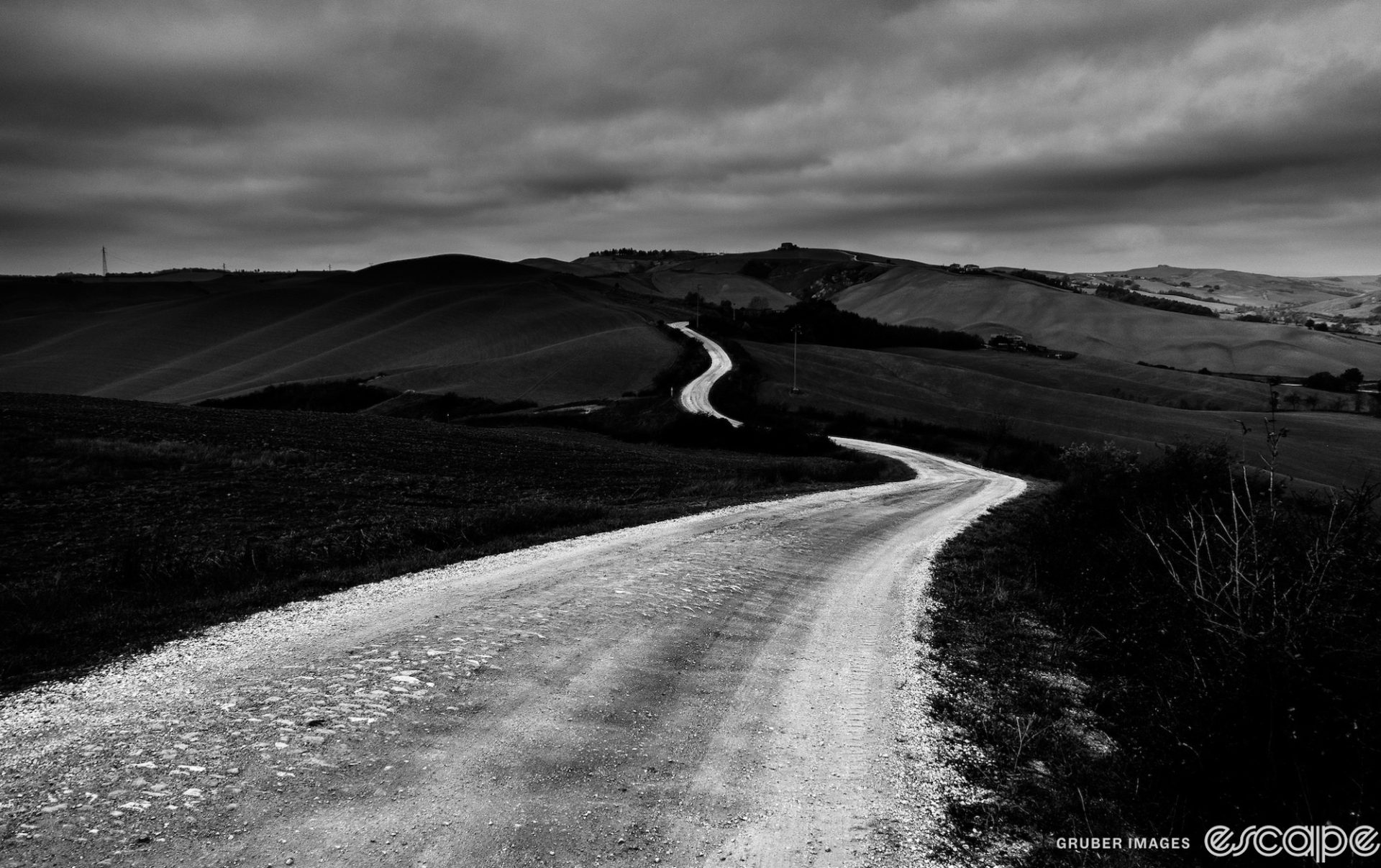 A black and white shot of a gravel road winding away in the distance over rolling hills. The fields are dark, as is the cloudy sky and it has a sense of foreboding.