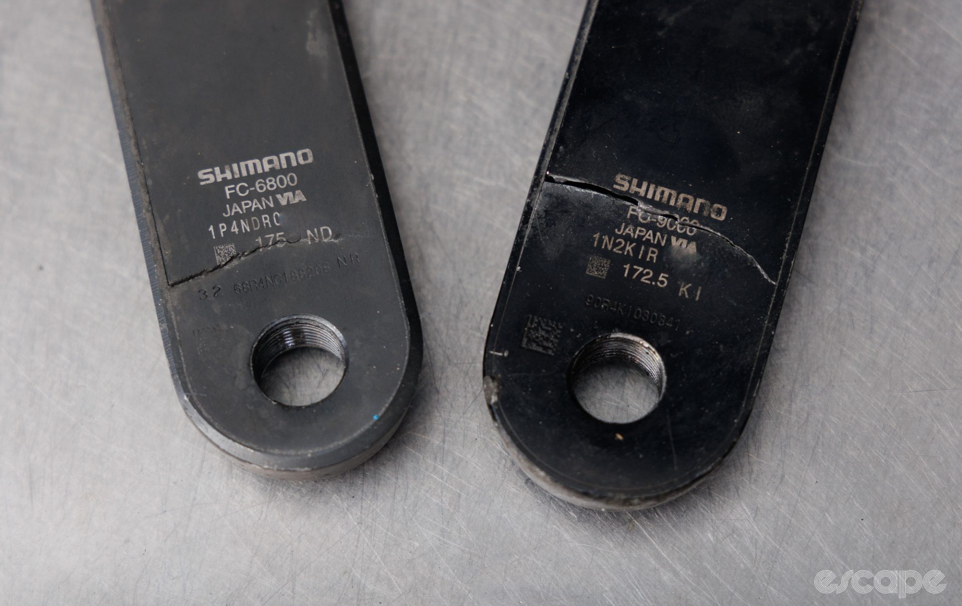 Two broken Shimano crankarms, shown from the inward facing side of the arms. Large, noticeable cracks spread across the arm in a perpendicular axis, and cross right through the manufacturing model and serial code stamp above the hole for the pedal spindle.