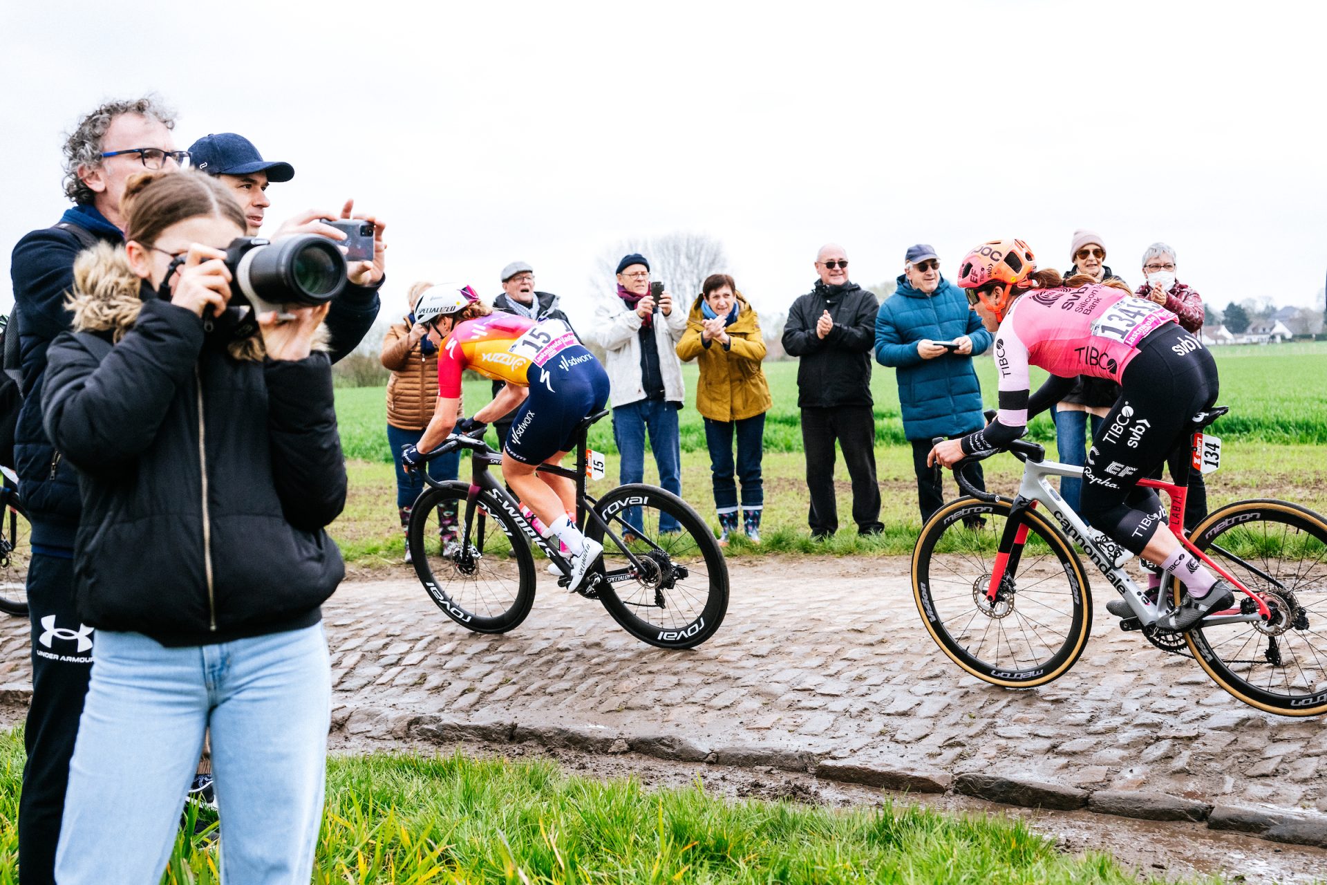 Fans and photographers gather on both sides of a narrow cobbled section at the 2023 Paris Roubaix Femmes. A string of riders fly past, navigating the slick cobbles.