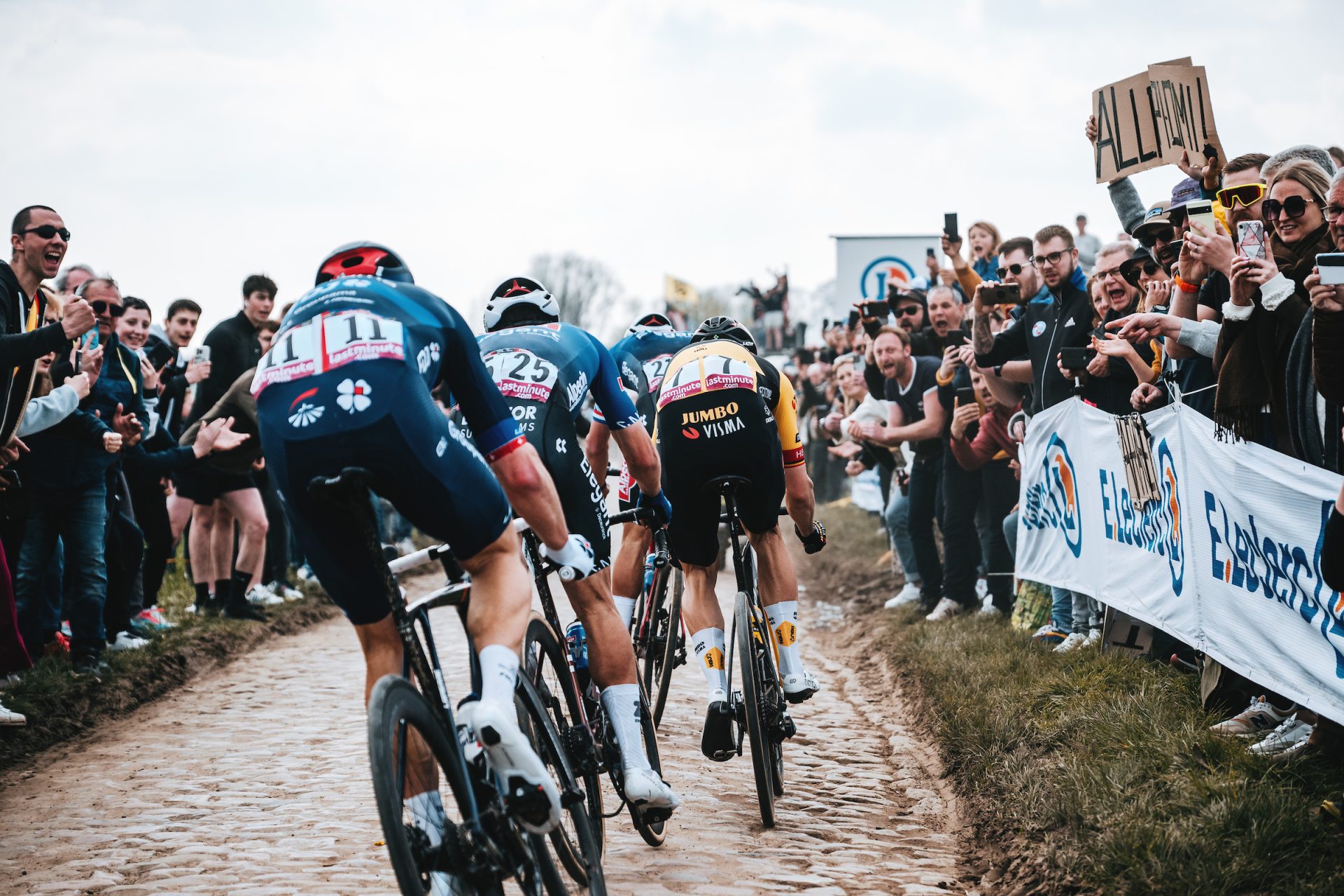From the perspective of a photographer on a moto, a tight grou of riders navigates the cobbles while flanked by rows of supporters at the edge of the road.