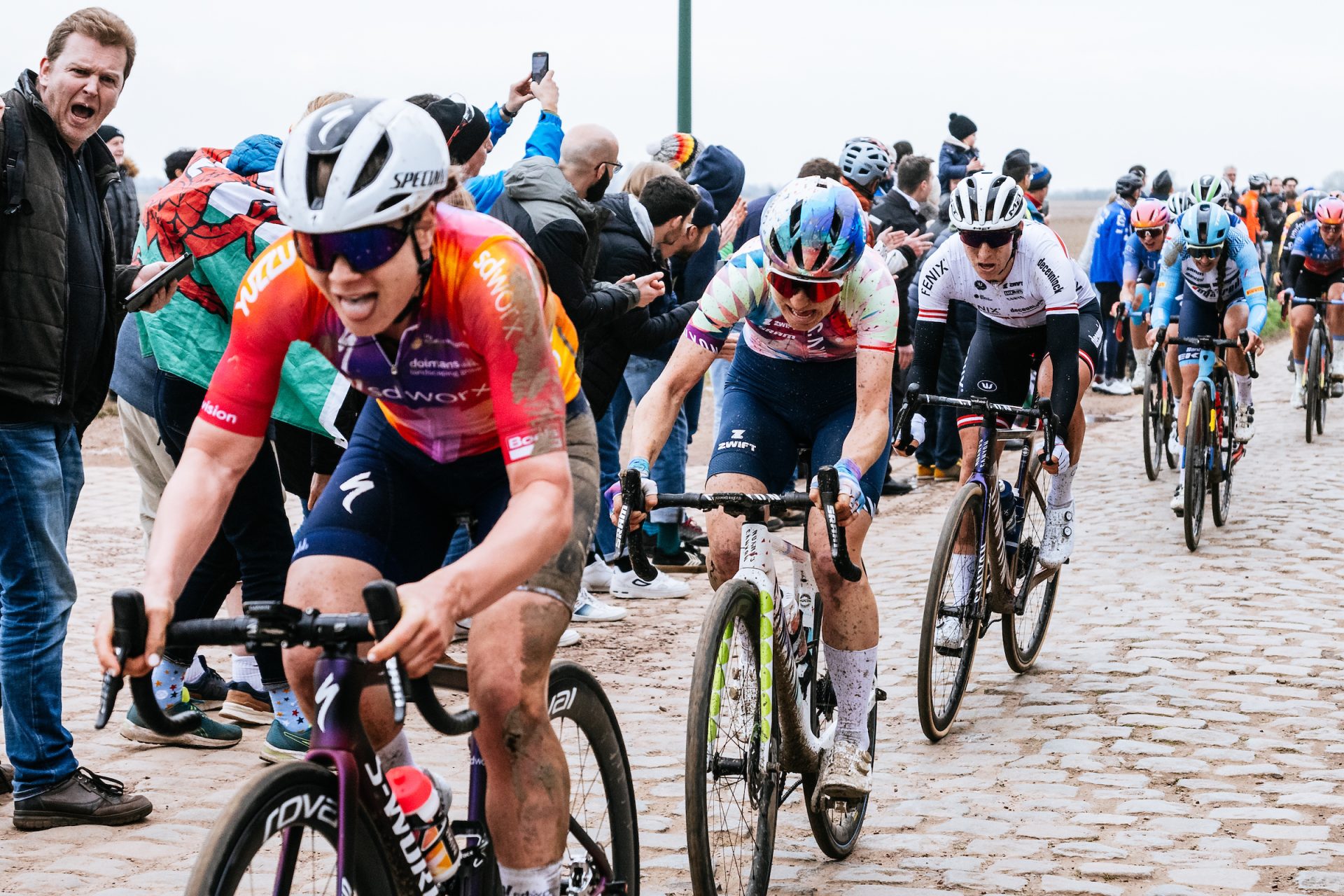 A tight shot of riders powering over the cobbles of the 2023 Paris Roubaix Femmes. The pack is stretched out, with multiple bike length gaps opening. Fans dressed in coats and rain jackets flank the side of the road.