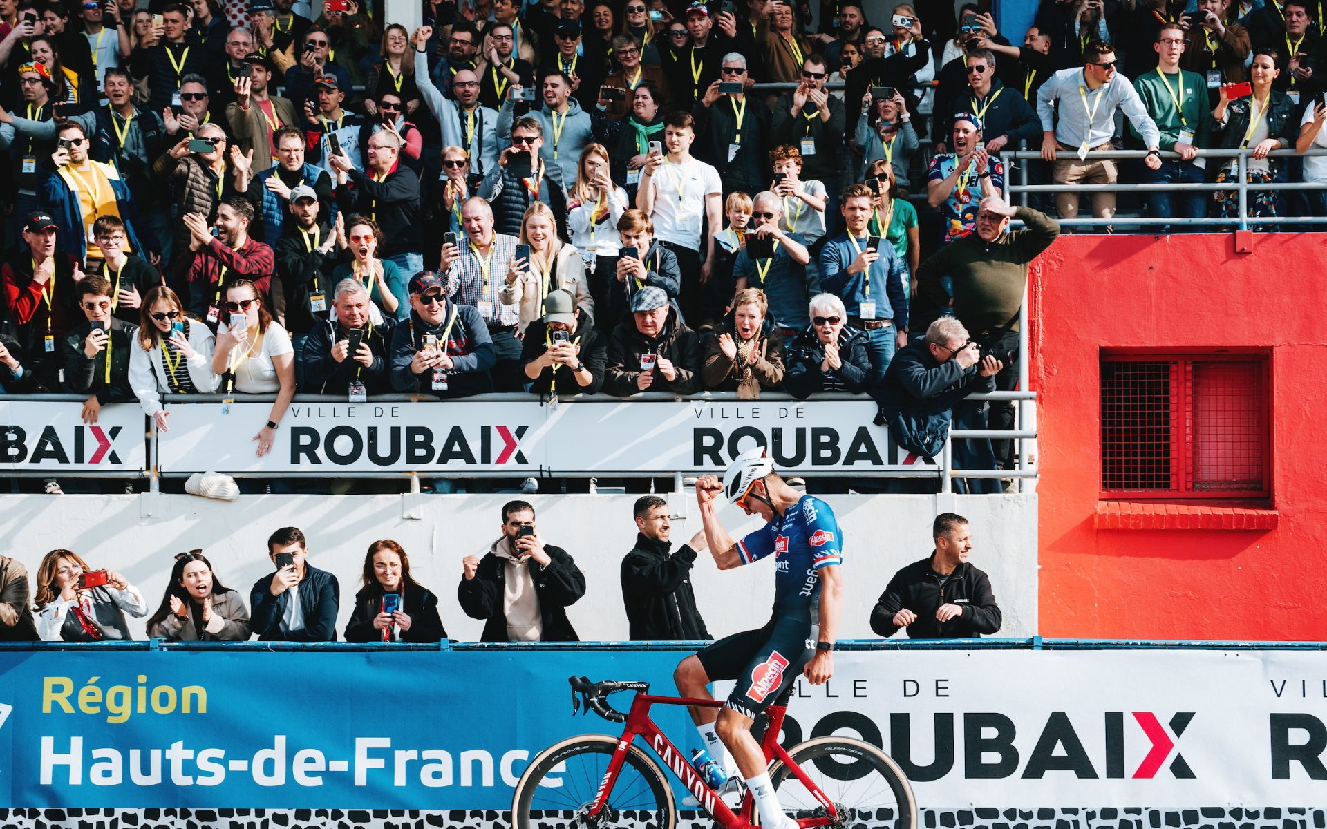 Mathieu van der Poel's victory salute, captured from the infield looking into the packed rafters. The blues, whites and reds of various sponsor banners and velodrome walls, as well as van der Poel's blue kit, white helmet and red bike, evoke the French and Dutch flags.