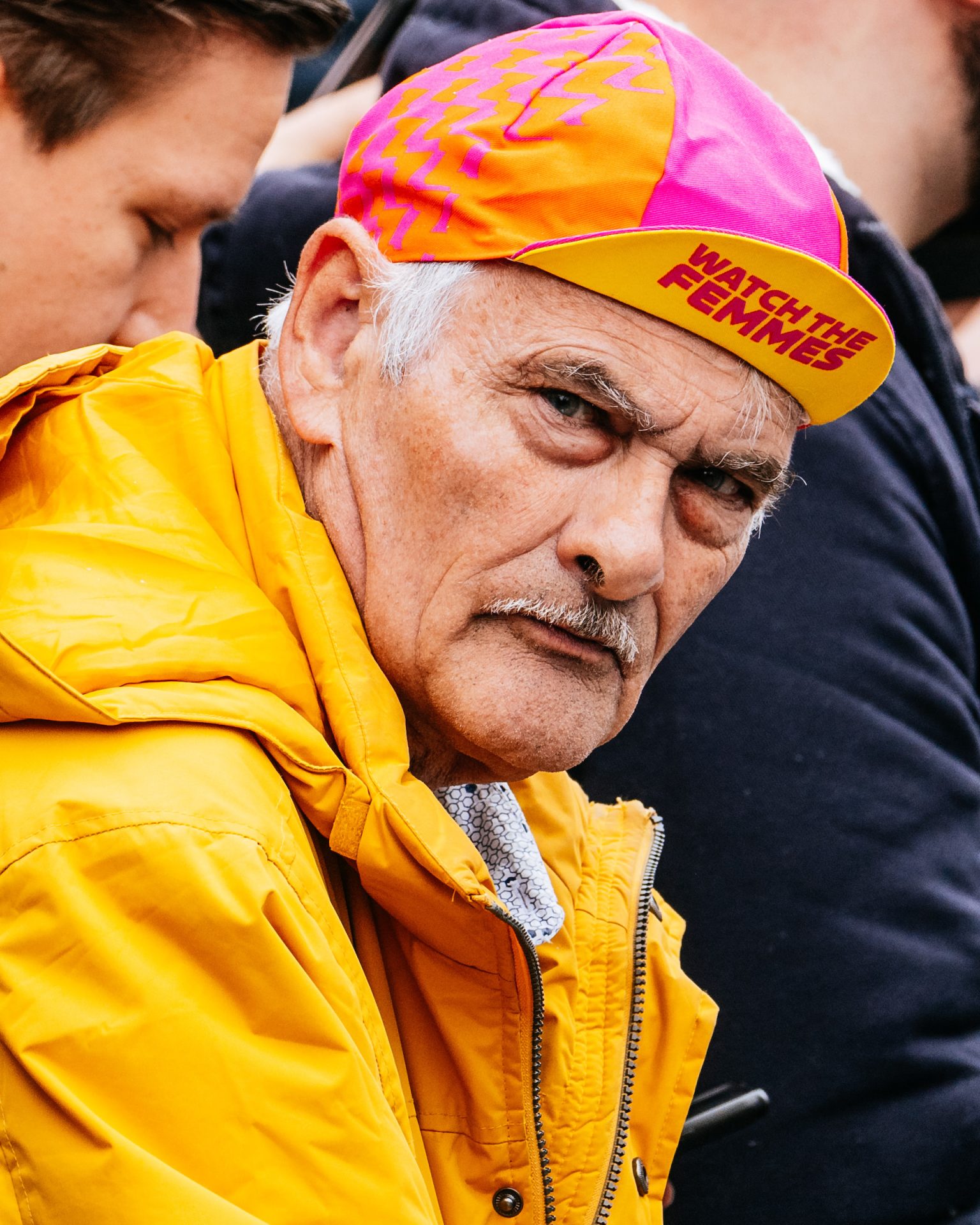 A close up of a supporter wearing a pink and yellow 'Watch the Femmes' cap. The man, with a thin moustache, wisps of white hair and a bright yellow rain jacket appears to be staring directly at the camera, his brow furled in deep concentration.