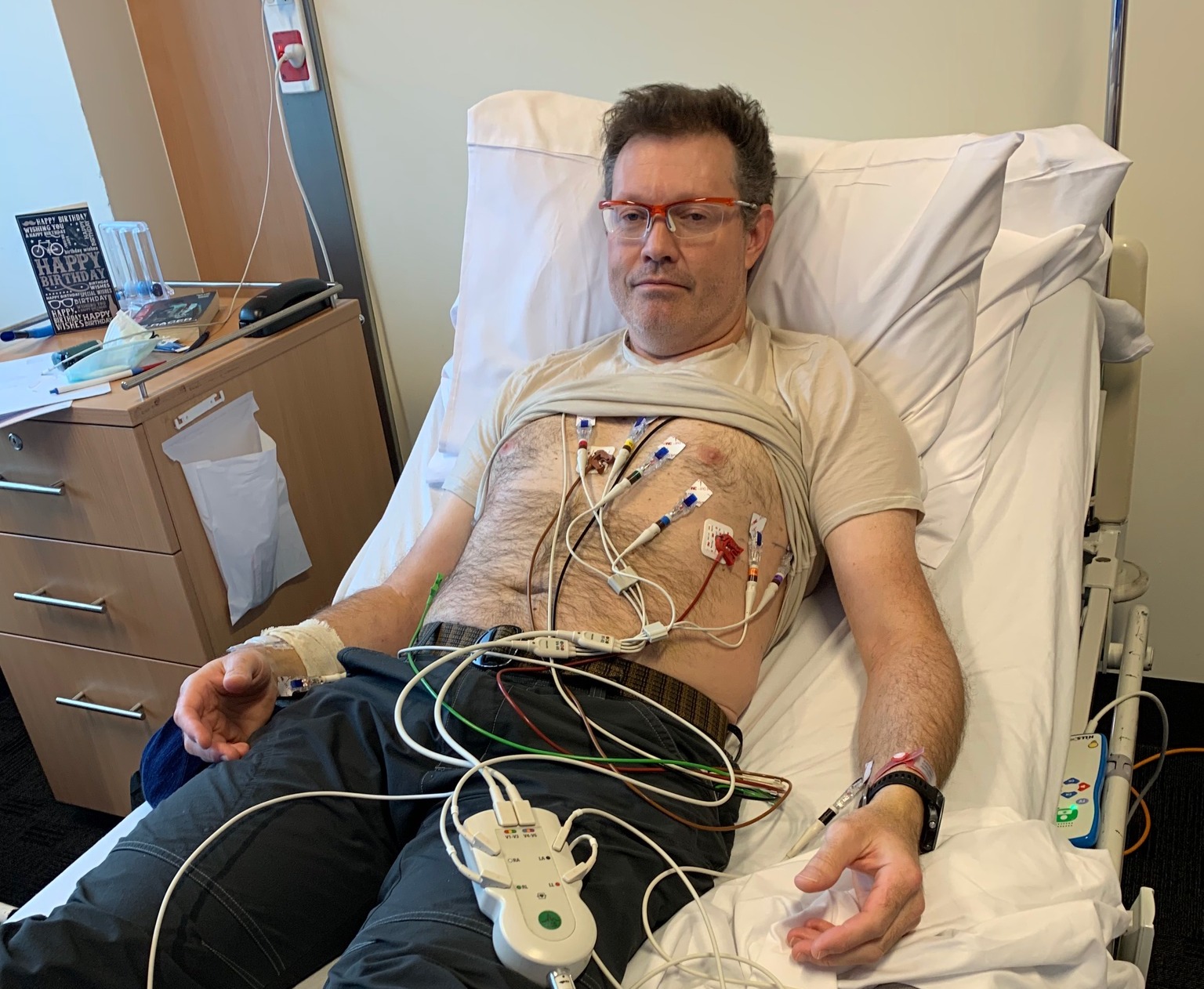 Jim Crumpler is reclined in a hospital bed with a multitude of wires attaches to his chest.