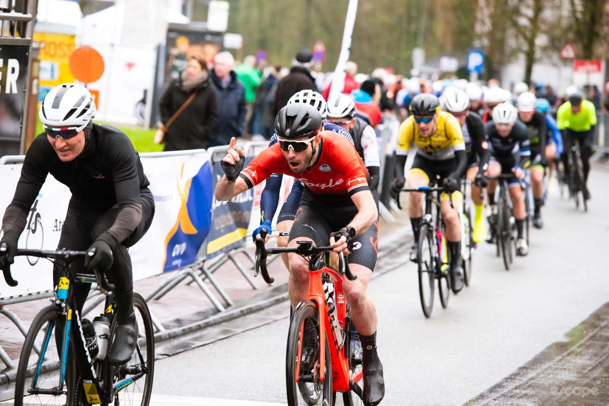 Ronan Mc Laughlin races in a Belgian kermesse. He's chasing a rider in all black, and has a hand raised as if he's about to tell him something.