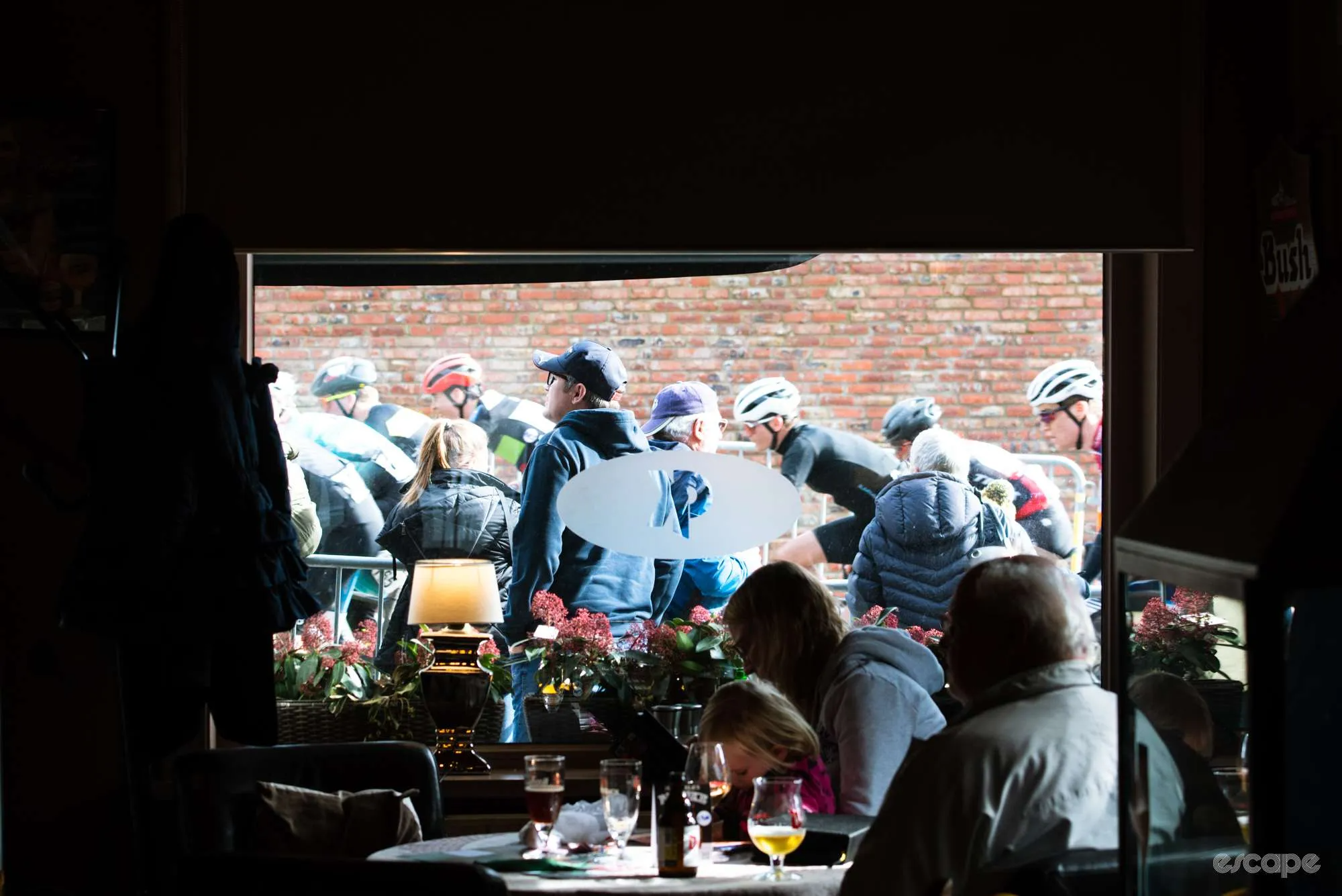 Fans watch a bike race from inside a restaurant, as riders pass by outside the window.