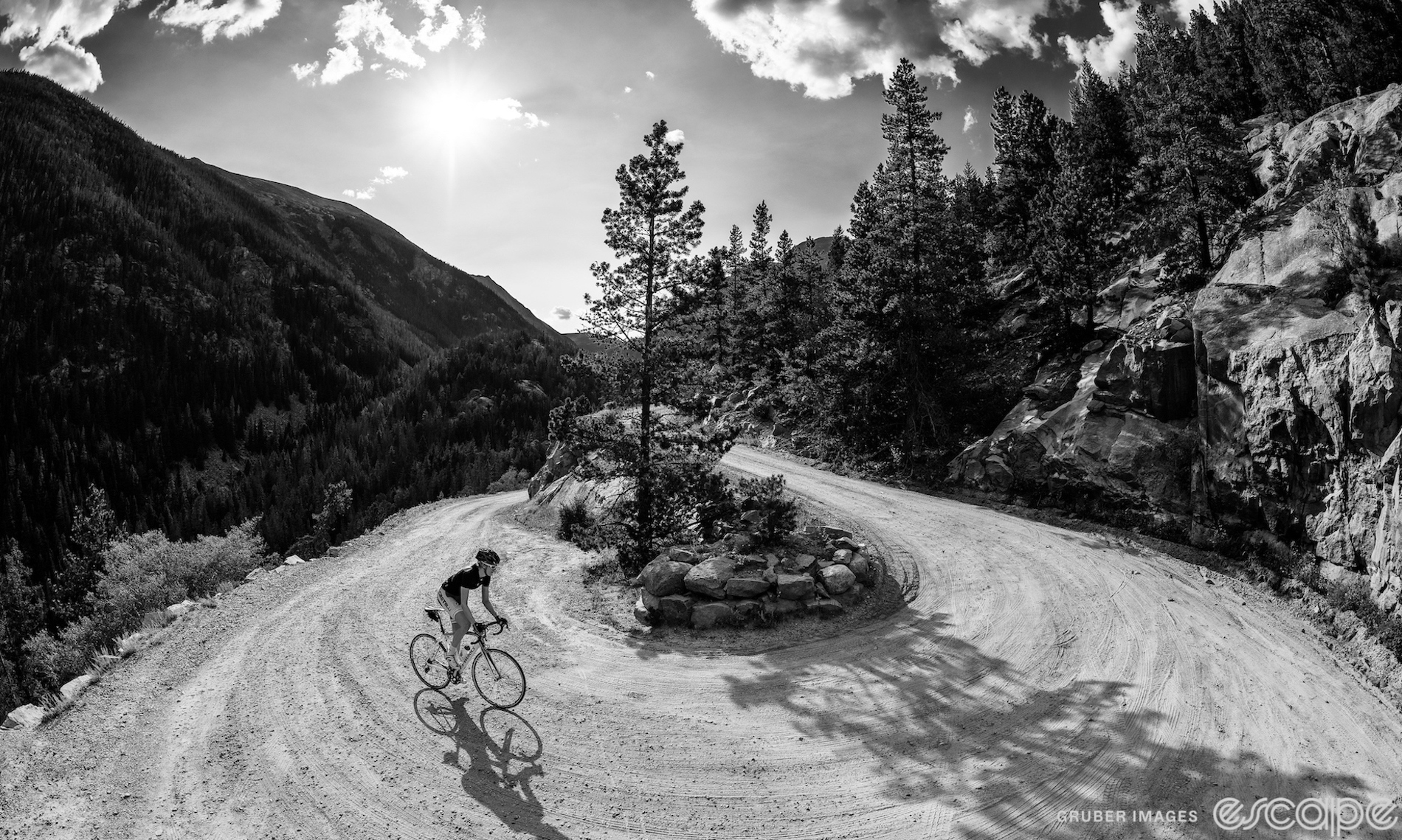 A lone cyclist rounds a hairpin on a dirt road. The black and white shot is a wide lens perspective from the outside apex of the corner, and the rider is out of the saddle as she rounds the corner with the sun shining on a massive ridgeline on the left side of the shot.