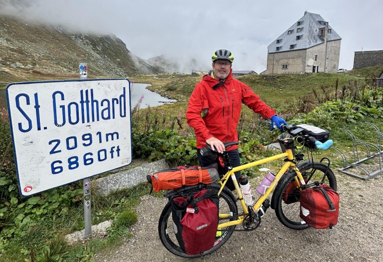 Jim Crumpler and his fully loaded bike stand next to a sign indicating the St Gotthard Pass.