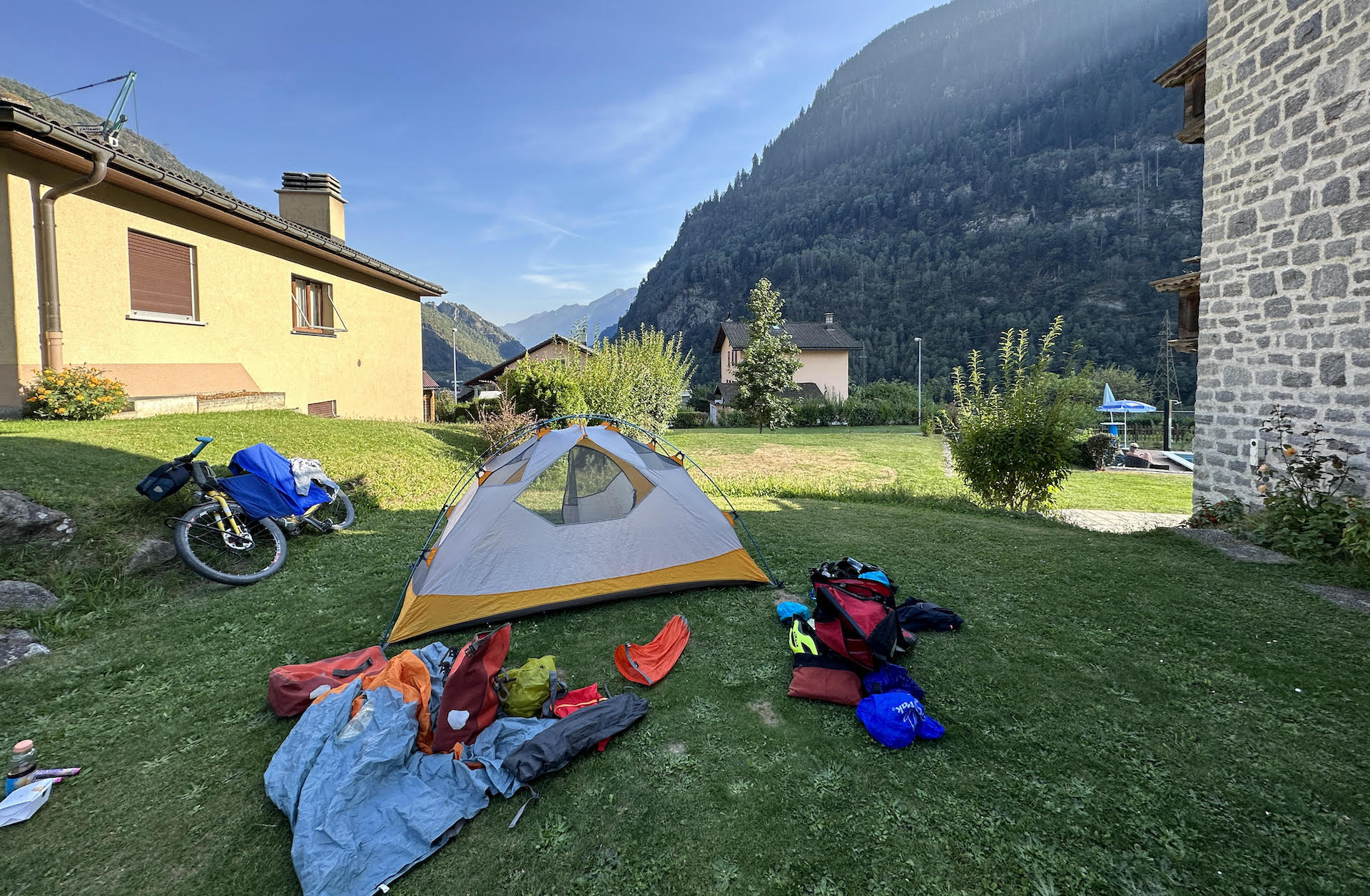 A tent is partly erected on some grass with Jim's bike and other gear strewn around it. There's a mountain in the background.