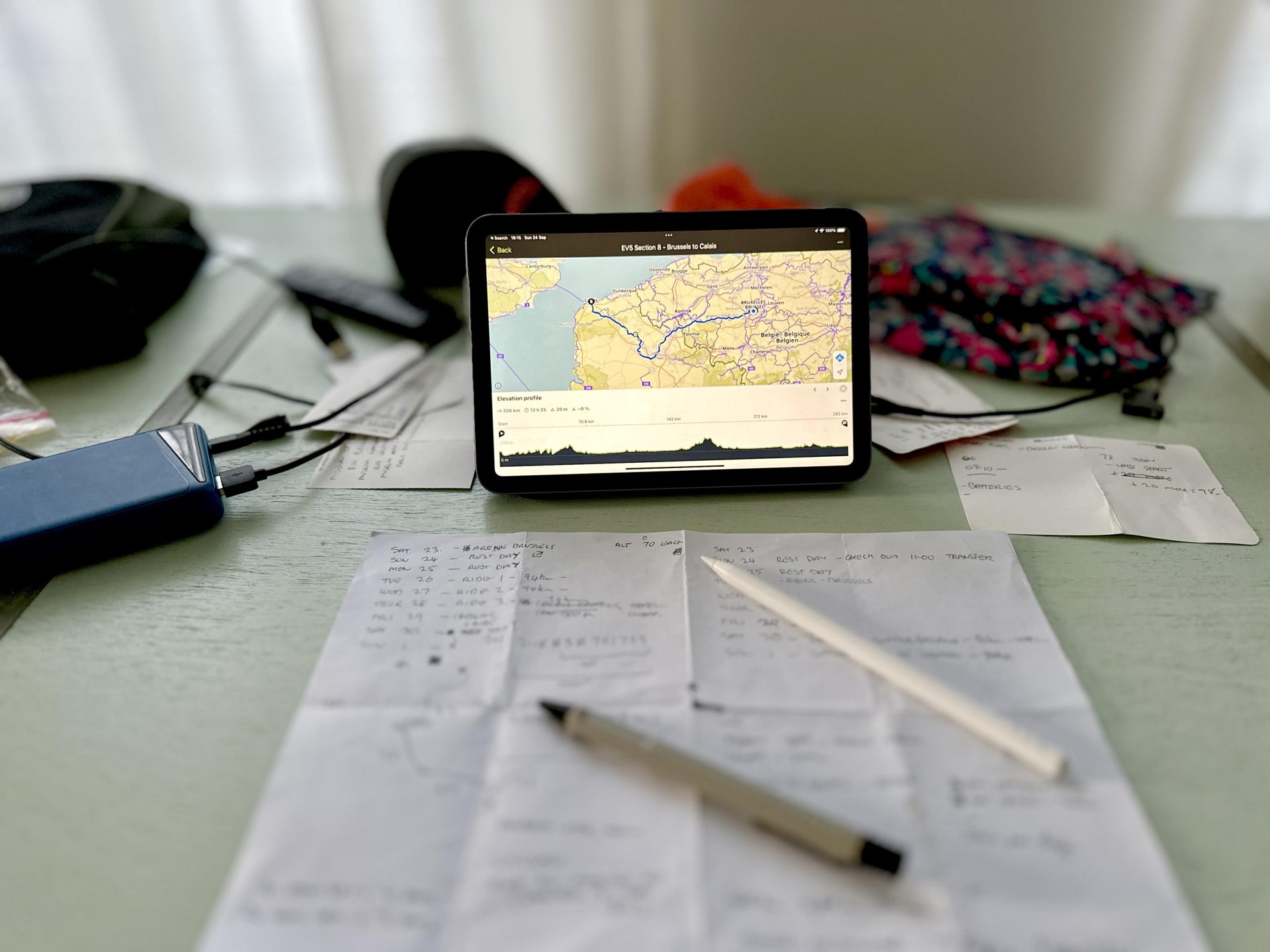 An iPad shows a bike route on screen while pens and paper little a table, with calculations about daily distances.