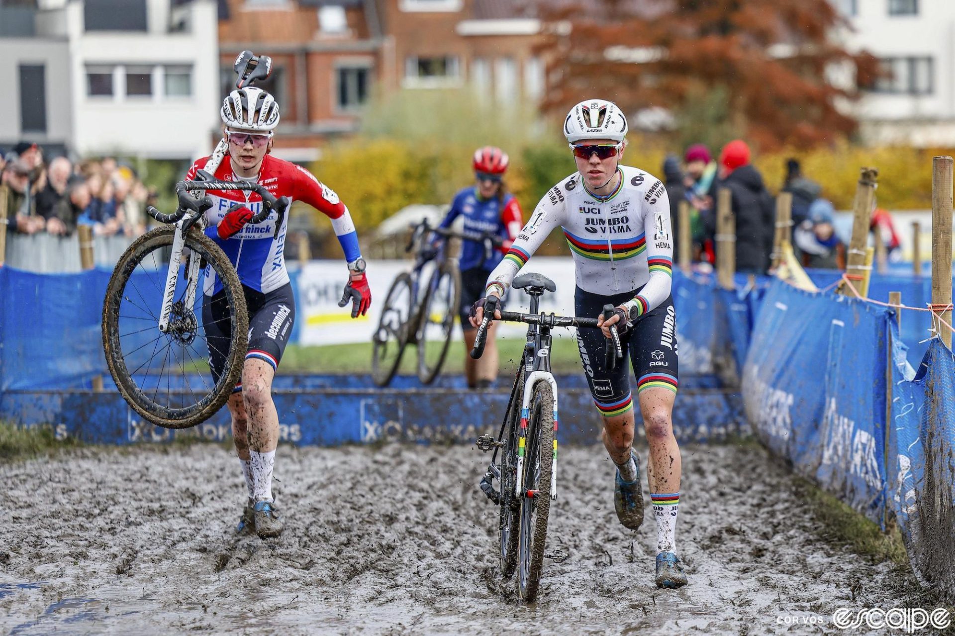 Fem van Empel runs her bike in the mud at a cyclocross race. She's just slightly ahead of rival Puck Pieterse, as both have crossed a set of barriers.