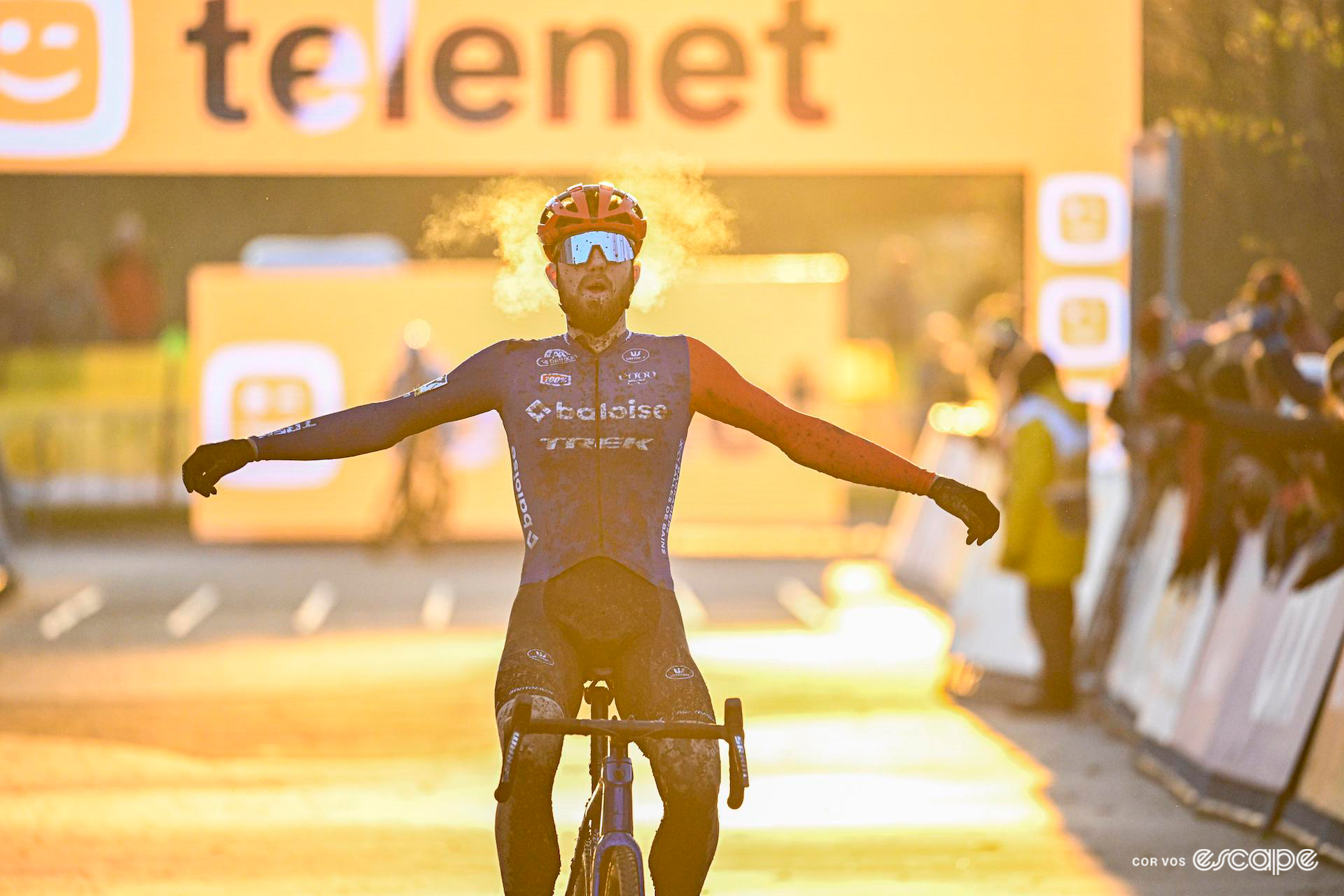 Joris Nieuwenhuis celebrates winning Cyclocross Superprestige Boom with his arms thrown wide, golden light illuminating his breath in the freezing cold temperatures.