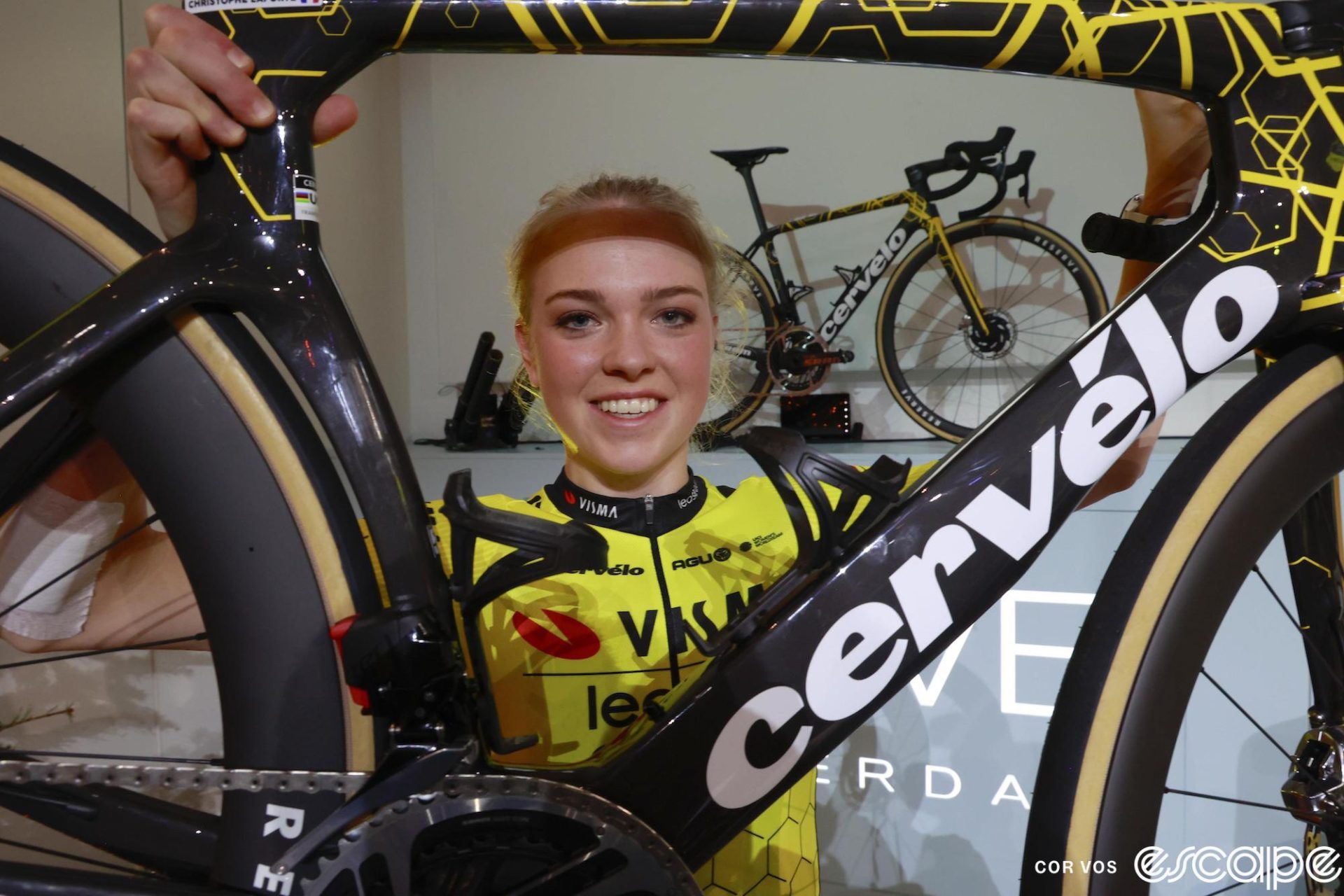 Fem van Empel holds up a Cervelo S5 road bike in team paint at the Visma-Lease a Bike team presentation. She's wearing the team's bright yellow and black kit and smiling as she's framed by the bike's main tubes.