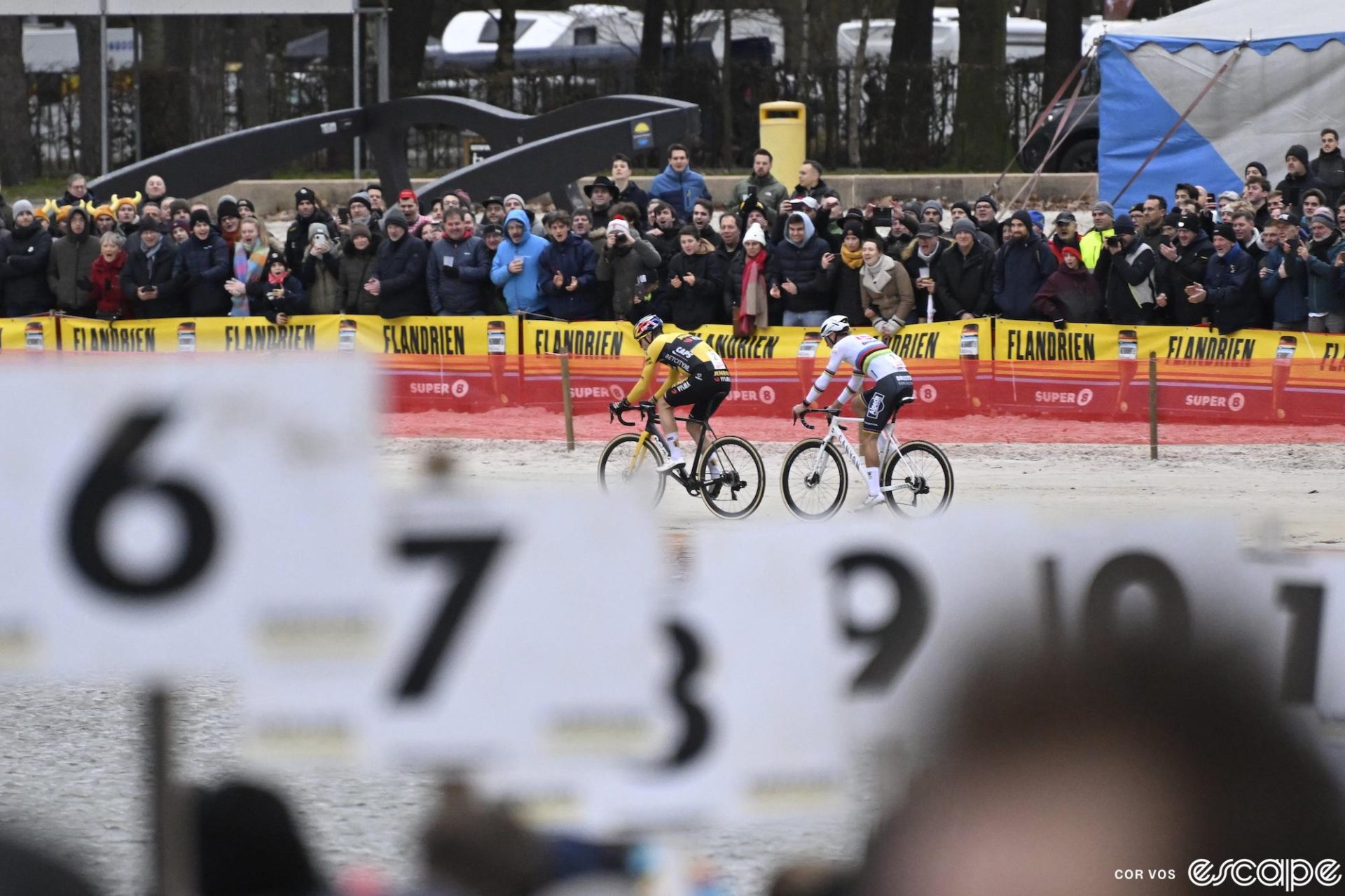 Wout van Aert leads Mathiu van der Poel in a sandy beach section at Zilvermeercross. The two are framed in front of a line of fans, looking out from the bike pit with numbered stalls.
