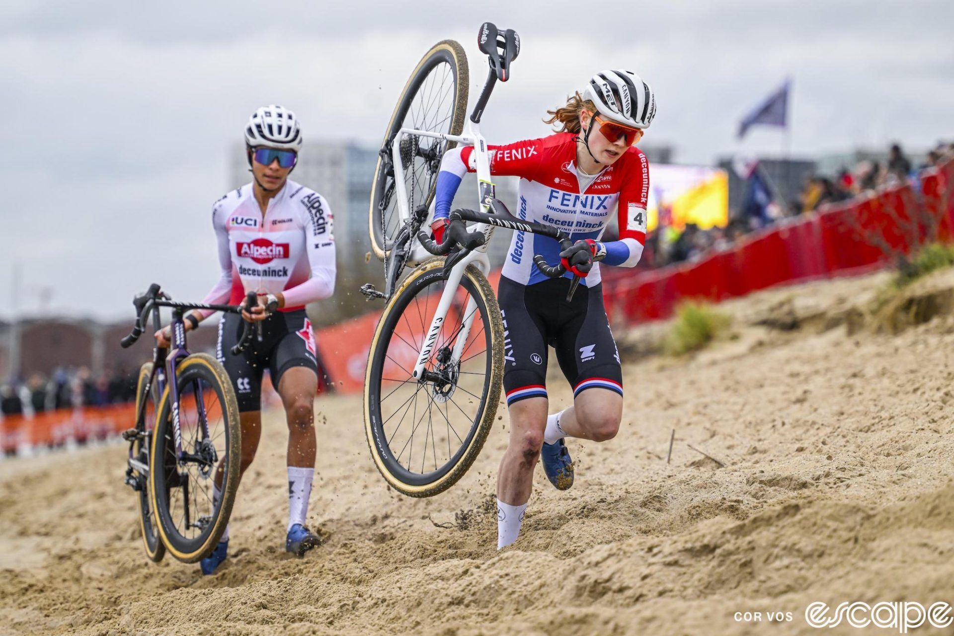 Puck Pieterse and Ceylin del Carmen Alvarado shoulder their bikes in the sand.