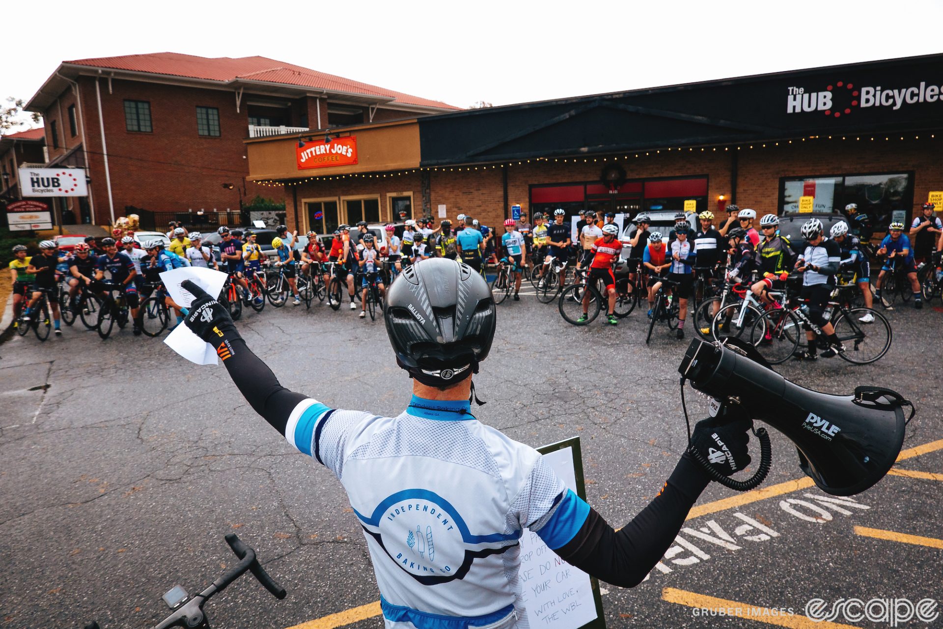 A cyclist and group ride leader addresses a pack of cyclists outside a bike shop before a group ride. He's holding a megaphone.