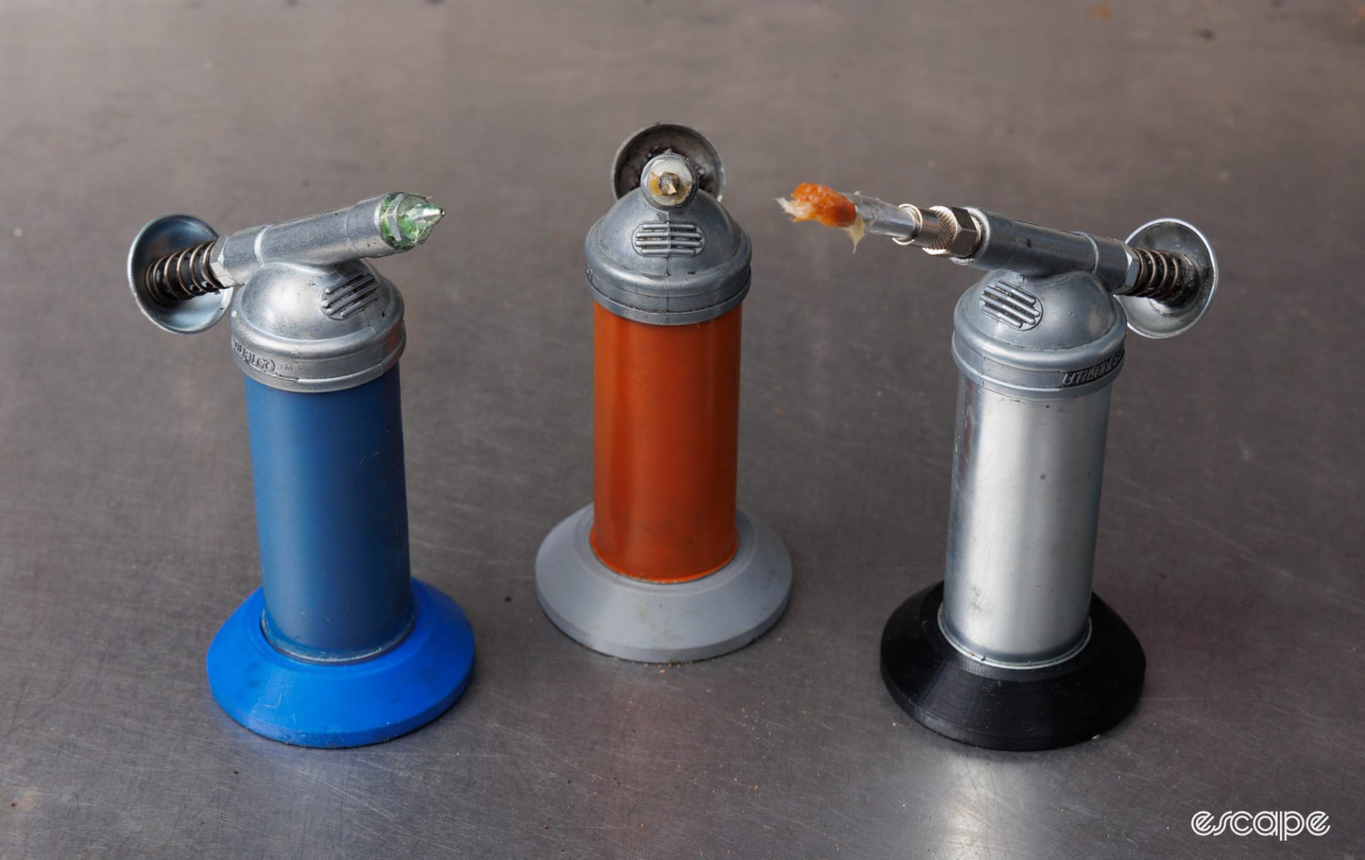 Three Dualco grease guns sitting on a benchtop. One is painted blue, another is painted orange, and the third one is raw. 